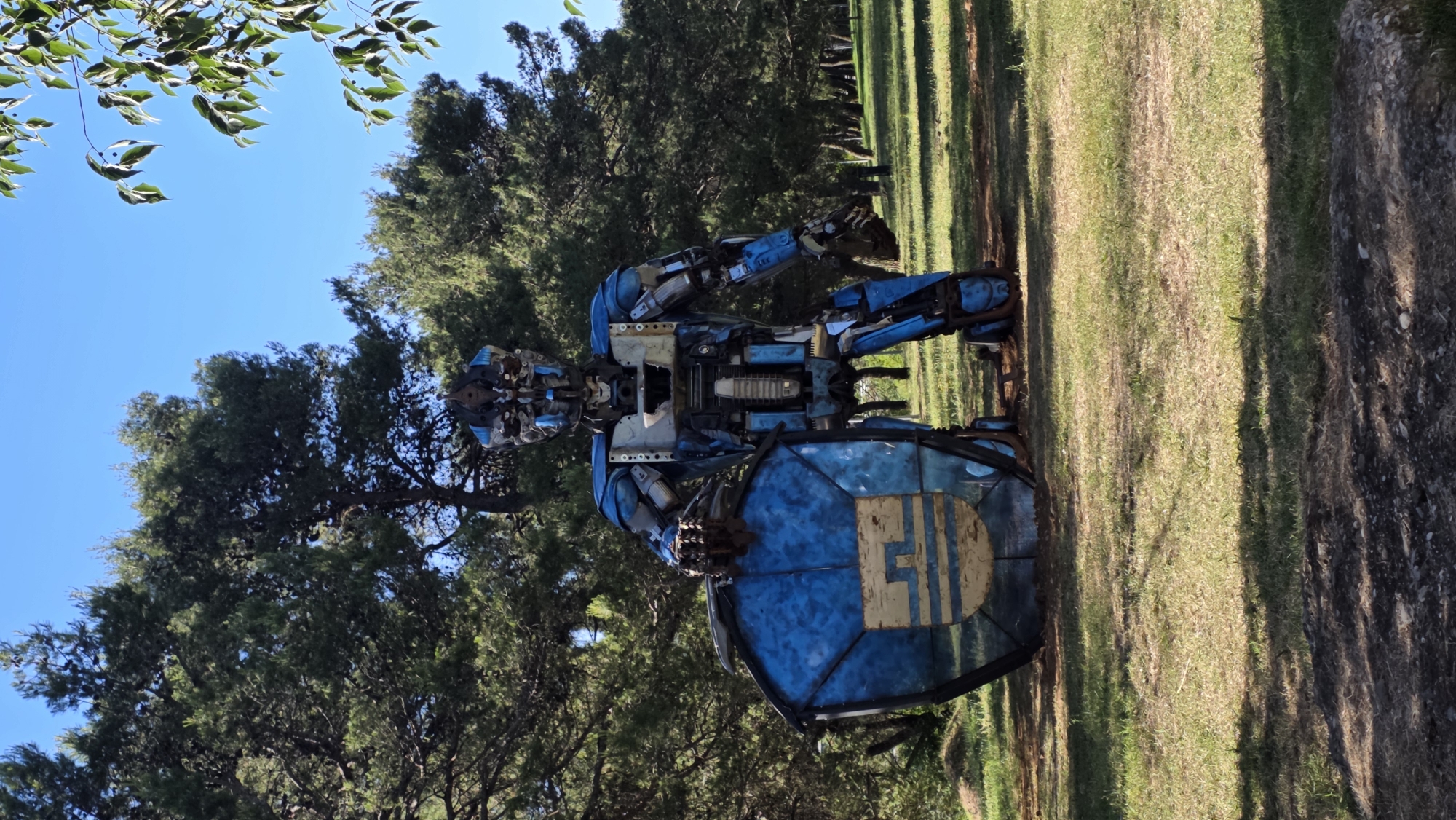 The image shows a blue robot sculpture standing on a grassy area in what appears to be a park setting. The robot is made of various metal parts. Trees are present in the background. The sky is visible, and the picture was taken outdoors on a sunny day. The location is likely in Podgorica, Montenegro.