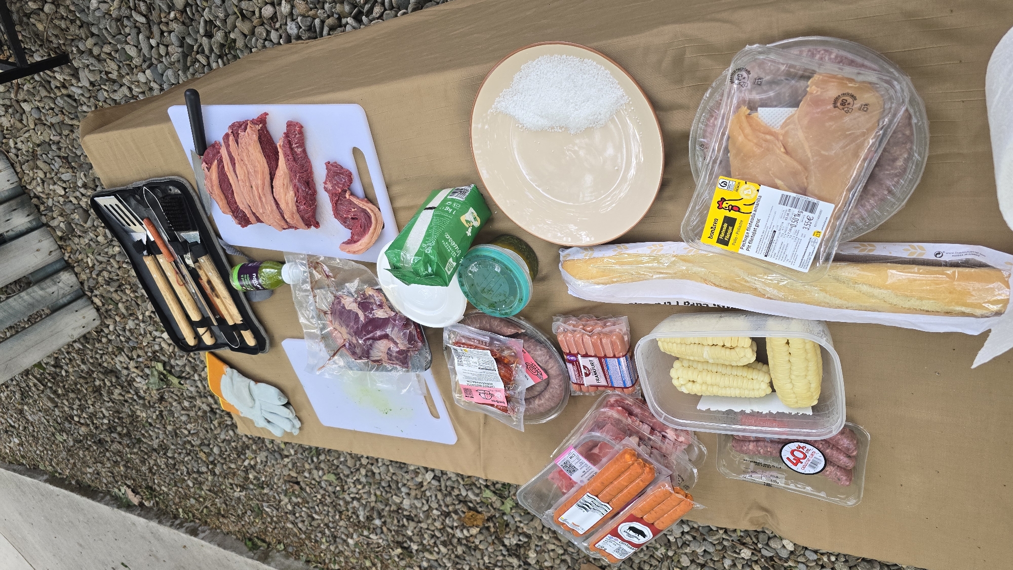 This image captures a spread of raw ingredients and grilling tools laid out on a brown cardboard surface, indicative of preparation for an outdoor barbecue or cookout, likely in Barcelona, Spain. The setting appears to be a gravel patio or backyard with some wooden elements visible on the left.
The main subjects are various cuts of meat and accompanying food items. On a white cutting board, several raw beef cuts, possibly picanha or similar, are neatly arranged alongside a large black-handled knife. A black case holds a set of wooden-handled grilling utensils, including a spatula, tongs, and a brush. Nearby, a small green bottle with "NATUR" visible on its label and a pair of white and orange gloves are present.
Other food items include a round plate piled with coarse salt. Packaged meats are prominent: bonÀrea chicken breast fillets ("Pechuga fileteada amarilla / Pit filetejat groc") with a label indicating "0.50 kg" and "3.55€," several packs of sausages including "FRANKFURT" and some marked "CADUCITAT CURTA" (short expiry), and ground meat packages, one displaying "40%." A green package marked "1 kg" is partially visible. A long baguette is wrapped in plastic, and a clear container holds cut corn on the cob. A roll of white paper towel is seen on the far right. The scene is illuminated by daylight, with no strong shadows or weather cues.