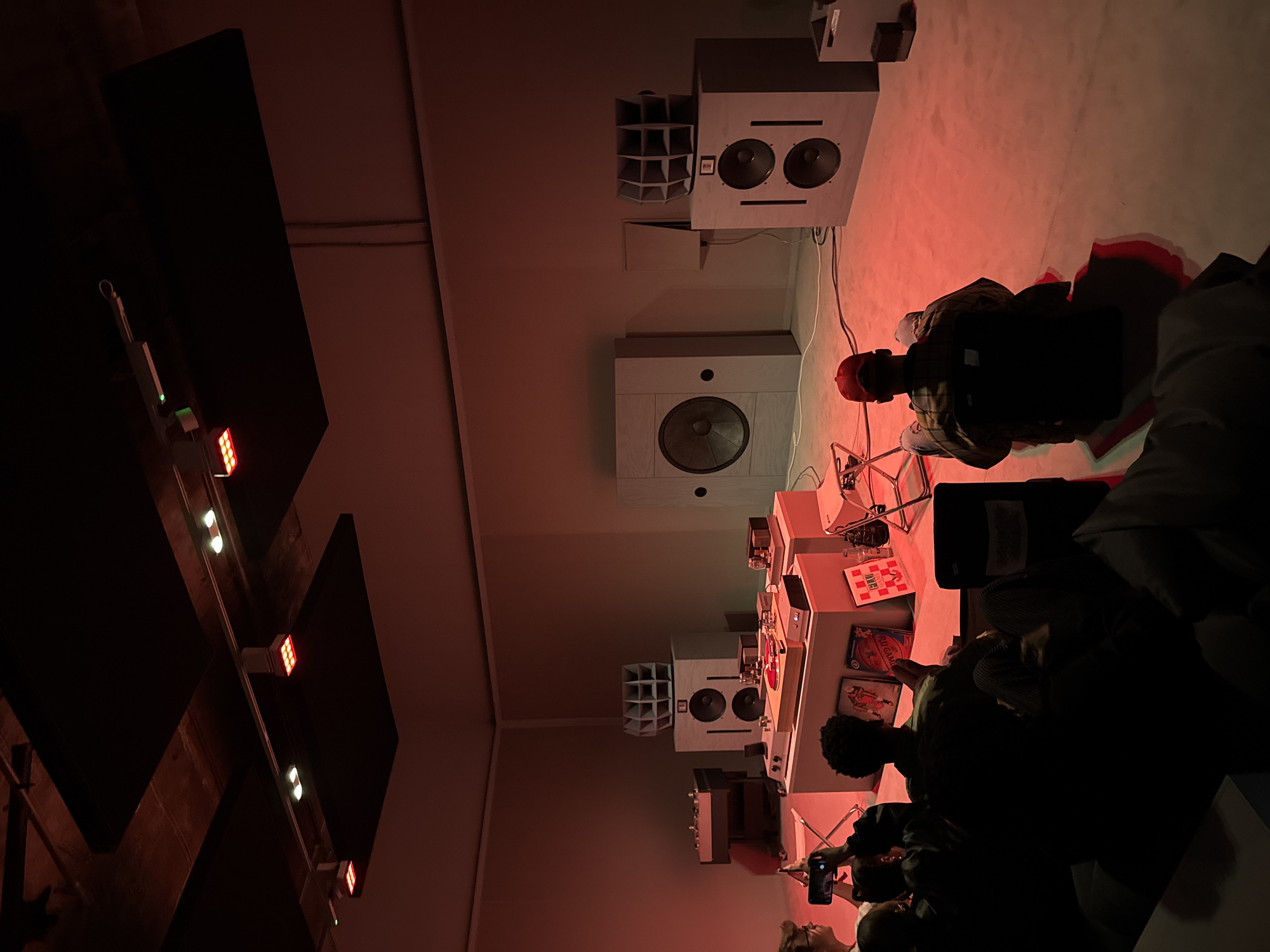 The scene depicts a gathering in what appears to be an audio or listening room. The room is illuminated with red light. Speakers are mounted on the wall. People are seated on the floor, listening to music from a setup consisting of record players and other audio equipment. The turntables are positioned on a low table surrounded by vinyl records. The location is suggestive of London, UK.