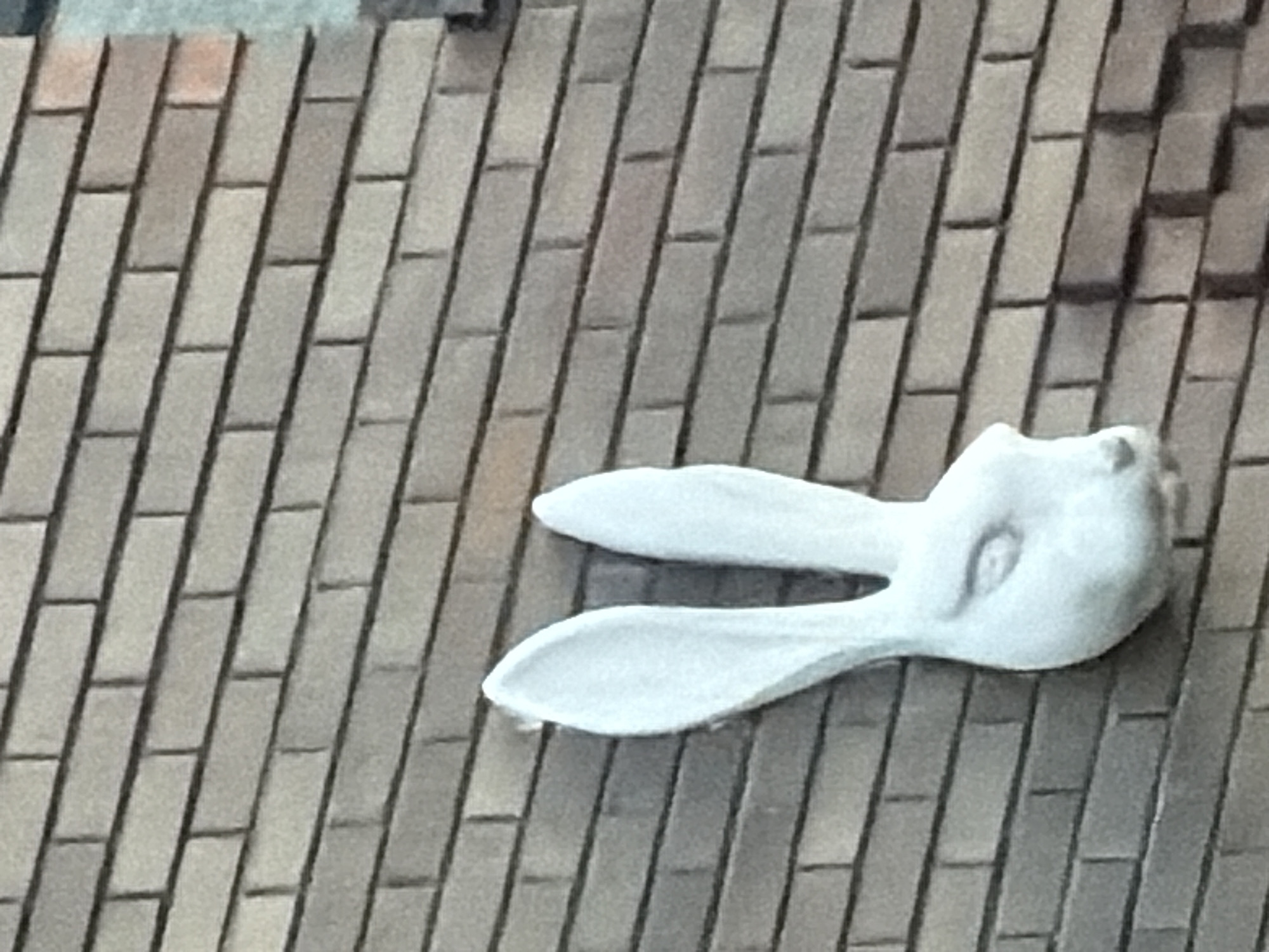 The image shows a white sculpture of a rabbit's head mounted on a brick wall. The sculpture is light-colored and appears to be made of plaster or a similar material. The rabbit's head faces to the right and has large, elongated ears that point upwards. Its eyes are indicated by simple indentations, and its nose and mouth are suggested by subtle contours. The brick wall is composed of rectangular bricks laid in a staggered pattern. The bricks are a muted brown and gray color, with visible mortar lines between them. The lighting suggests it is daytime. The specific location in Moscow, Russia, cannot be determined from the provided image.