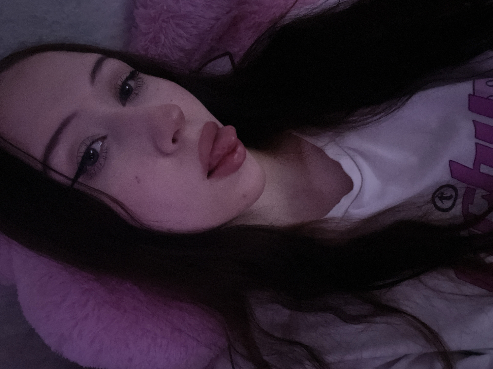 A young woman with dark hair and heavy eyeliner is lying down, looking at the camera. She is wearing a white t-shirt and has her tongue sticking out. She is leaning against a plush pink surface. The image is dimly lit and appears to be taken indoors. There is no information in the image to suggest a location in Votkinsk, Russia.