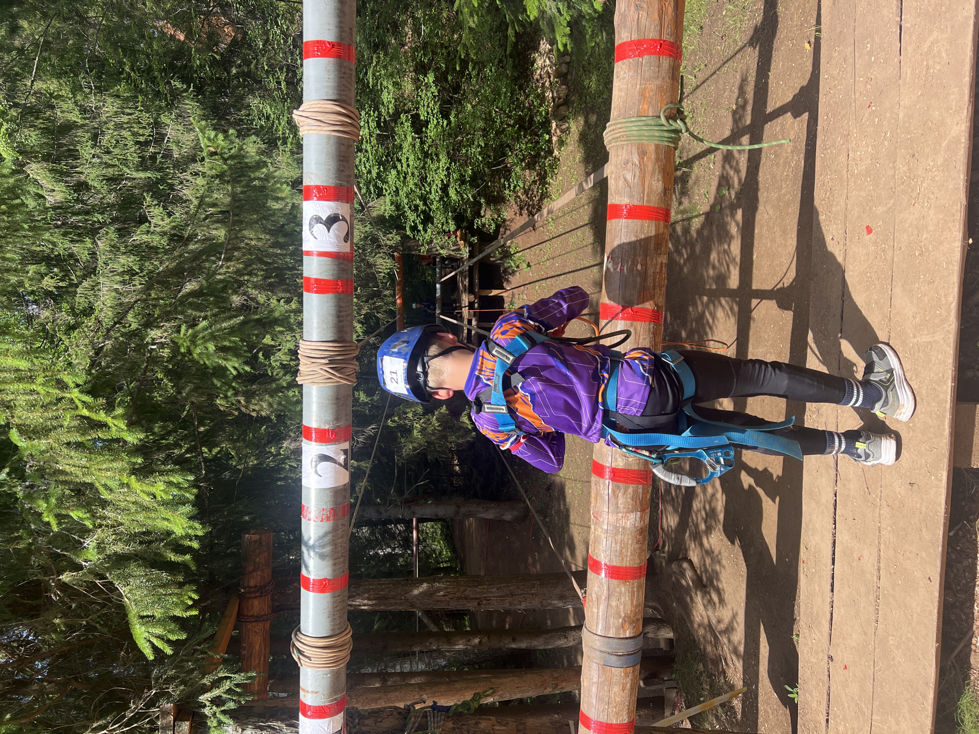 A young person, wearing a blue helmet and a purple and orange harness, is participating in an outdoor ropes course. They are clipped into a safety line and are positioned between two wooden poles that are part of the course. The poles are wrapped with rope and have red tape marking them at intervals. The young person is wearing dark leggings and athletic shoes. The background consists of lush green trees, indicating a forest setting. Sunlight is casting shadows on the wooden planks of the course, suggesting it is daytime. The scene appears to be in a recreational area designed for adventure activities. On one of the poles, there is a white label with the number "3" inside a black heart shape, and another label with the number "2". Another pole has the number "21" visible on a white sticker on the helmet. The overall impression is one of an active and adventurous outing.