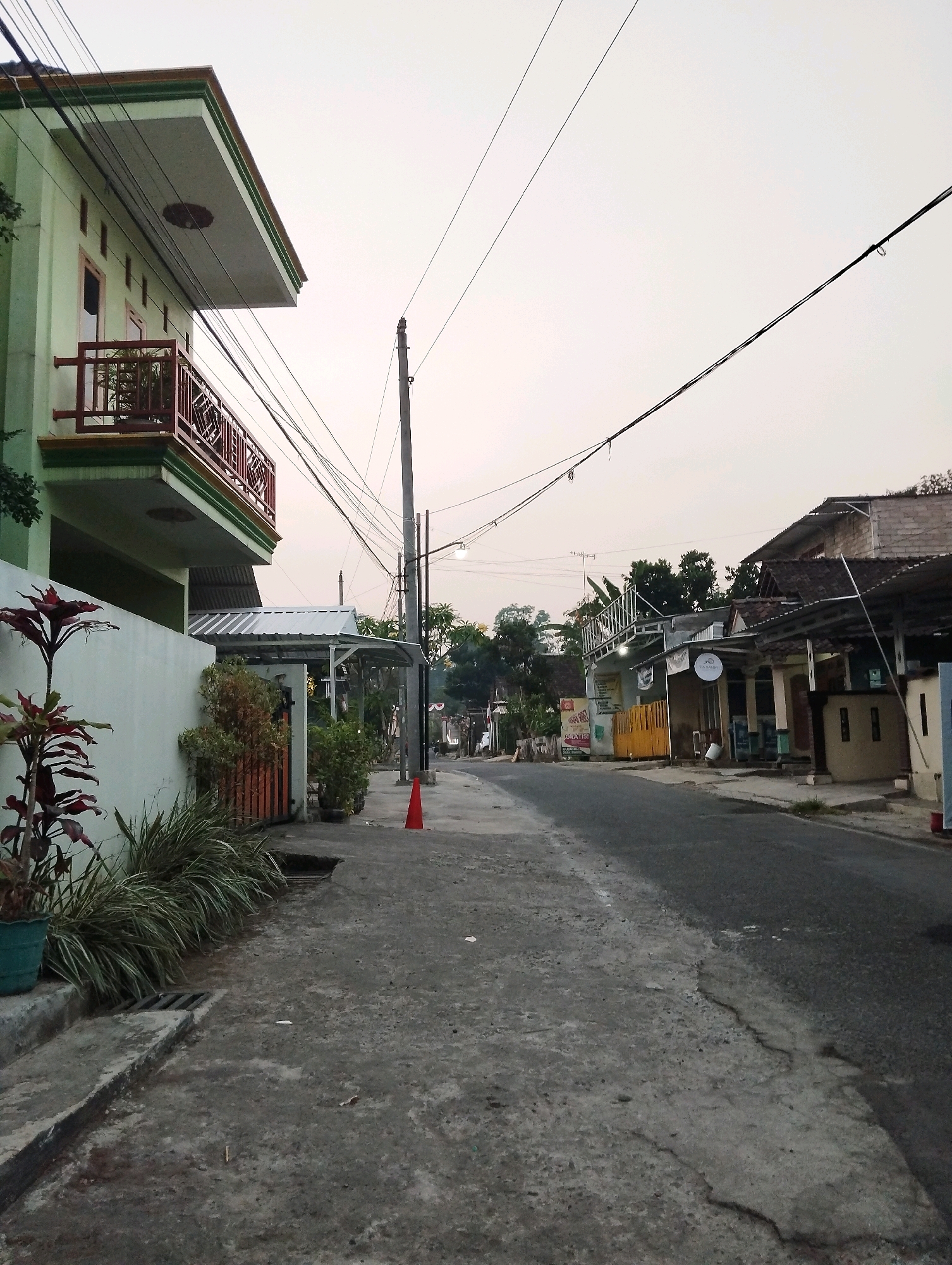 This is an eye-level, medium shot captured during the day in a residential area of Salatiga, Indonesia. The scene depicts a paved road with a sidewalk on the left, lined with houses and vegetation. A light green house with a red-railed balcony is prominent on the left, and a taller building with a multi-tiered roof is visible further down the street on the right. Power lines crisscross overhead, supported by utility poles. Various plants, including a tall plant in a green pot, are situated along the sidewalk. A bright orange traffic cone is placed on the sidewalk in the middle ground. The sky is overcast, suggesting a cloudy day. The overall impression is of a typical street in a suburban or semi-urban Indonesian neighborhood.