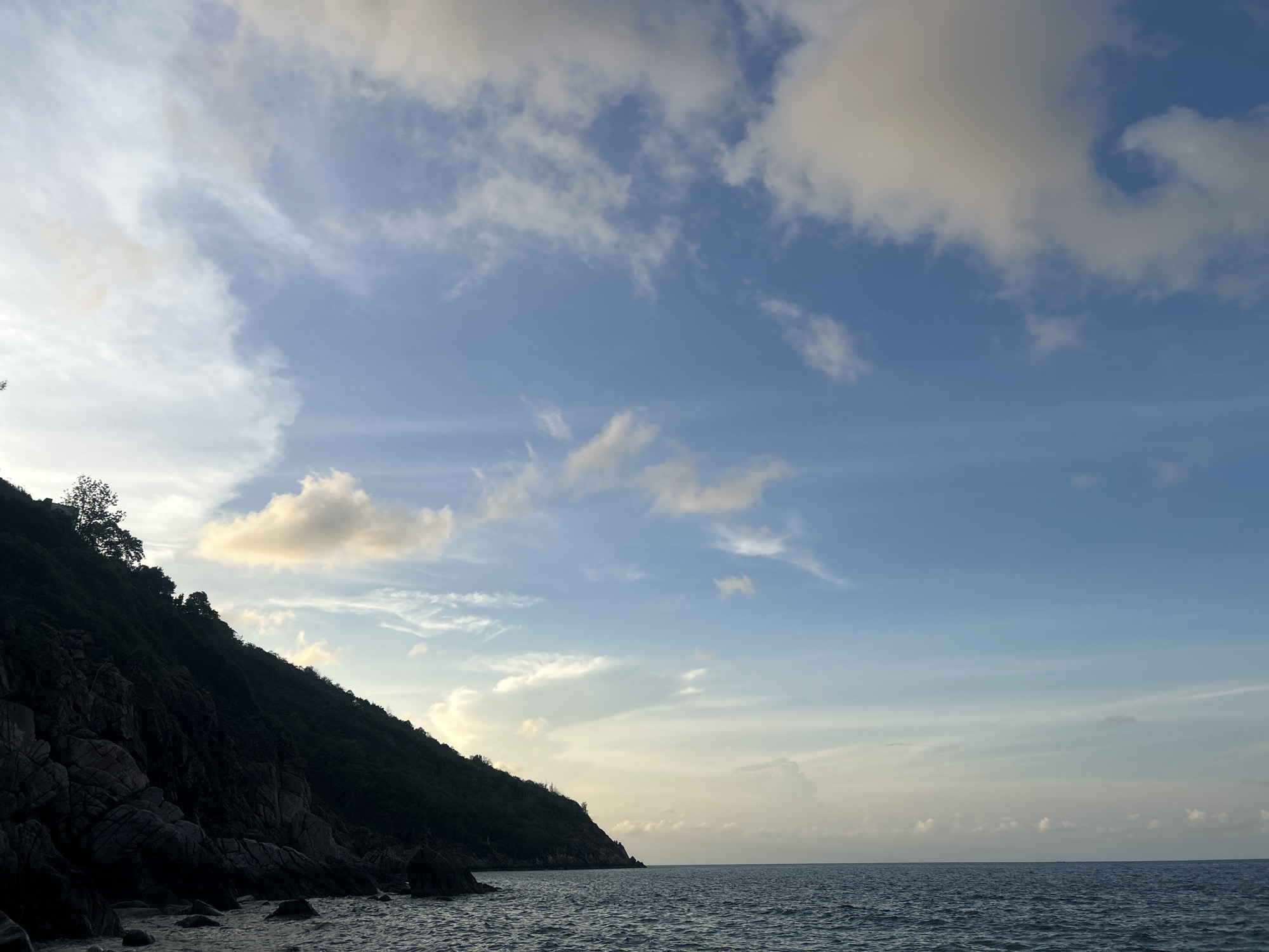 The image shows a coastal scene in Baan Chaloklum, Thailand. A steep, dark green, rocky hillside occupies the left side of the frame, with some trees at the top. The lower part of the hill transitions to a rocky beach with ocean water. The ocean, reflecting the sky, stretches horizontally across the bottom half of the image. The sky is predominantly blue, with scattered white clouds, some tinged with yellow. The horizon is visible in the distance. No people are visible.