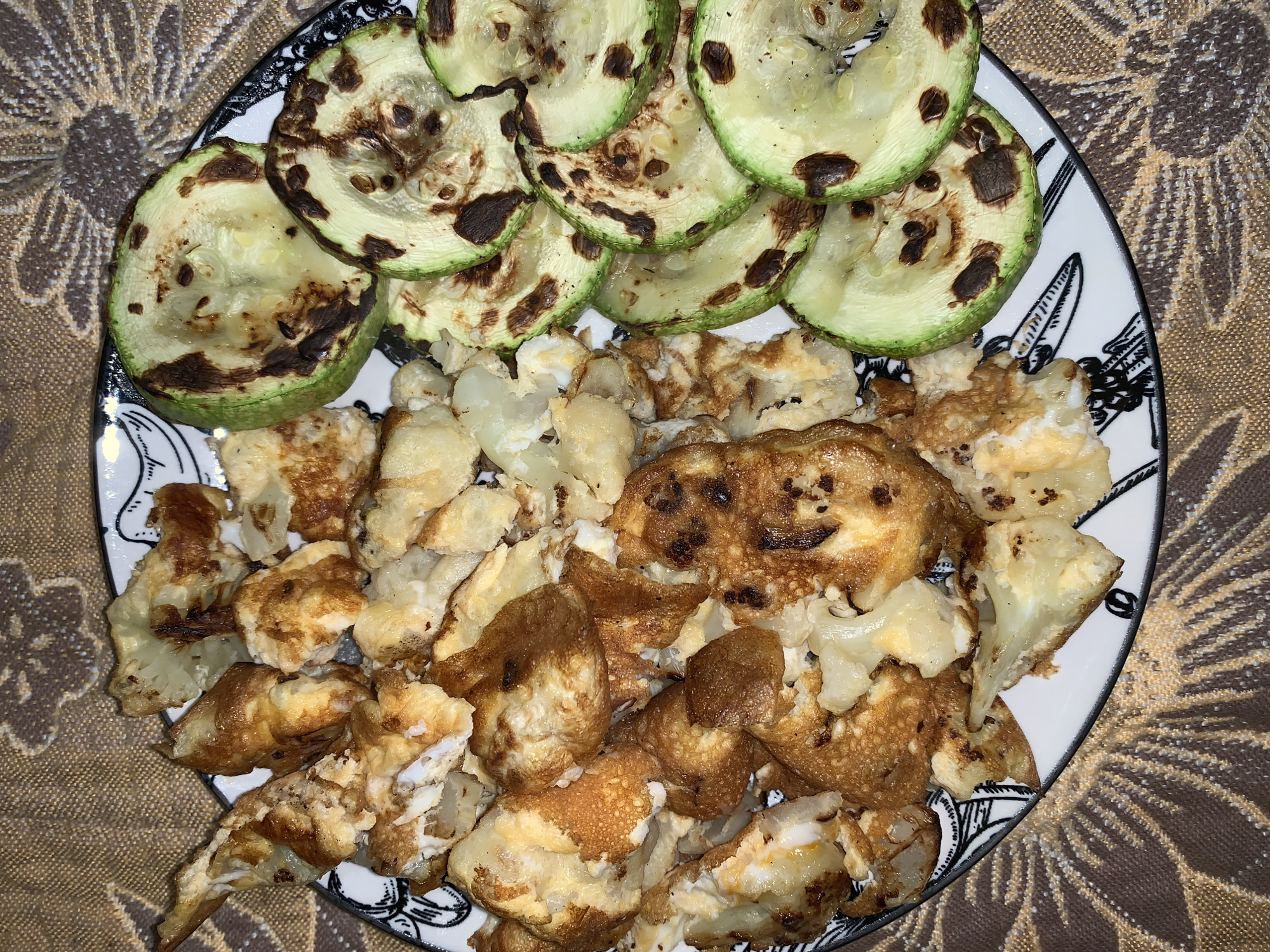 An overhead shot captures a meal consisting of grilled zucchini slices and fried cauliflower florets served on a decorative plate, set against a patterned brown tablecloth.
The top half of the plate is filled with thin, round slices of zucchini, characterized by their light green color and prominent, dark brown grill marks. These marks indicate they were cooked on a grill or griddle, suggesting a charred, smoky flavor.
Below the zucchini, the plate holds numerous irregular pieces of cauliflower or a similar white vegetable. Many of these pieces are coated and fried, showing golden-brown and crispy surfaces, while others are lighter in color, revealing the natural white of the vegetable. The texture appears varied, with some parts being soft and others crunchy.
The food is presented on a white ceramic plate that features a distinctive black and white decorative rim. The pattern on the rim includes abstract or stylized elements, possibly resembling cutlery or intricate floral designs. The plate rests on a brown fabric surface, which appears to be a tablecloth or placemat, adorned with a lighter brown or beige floral, sunburst-like pattern.
The lighting in the image suggests an indoor setting. There are no people visible, nor any discernible text. The arrangement of the food suggests a prepared meal ready for consumption.