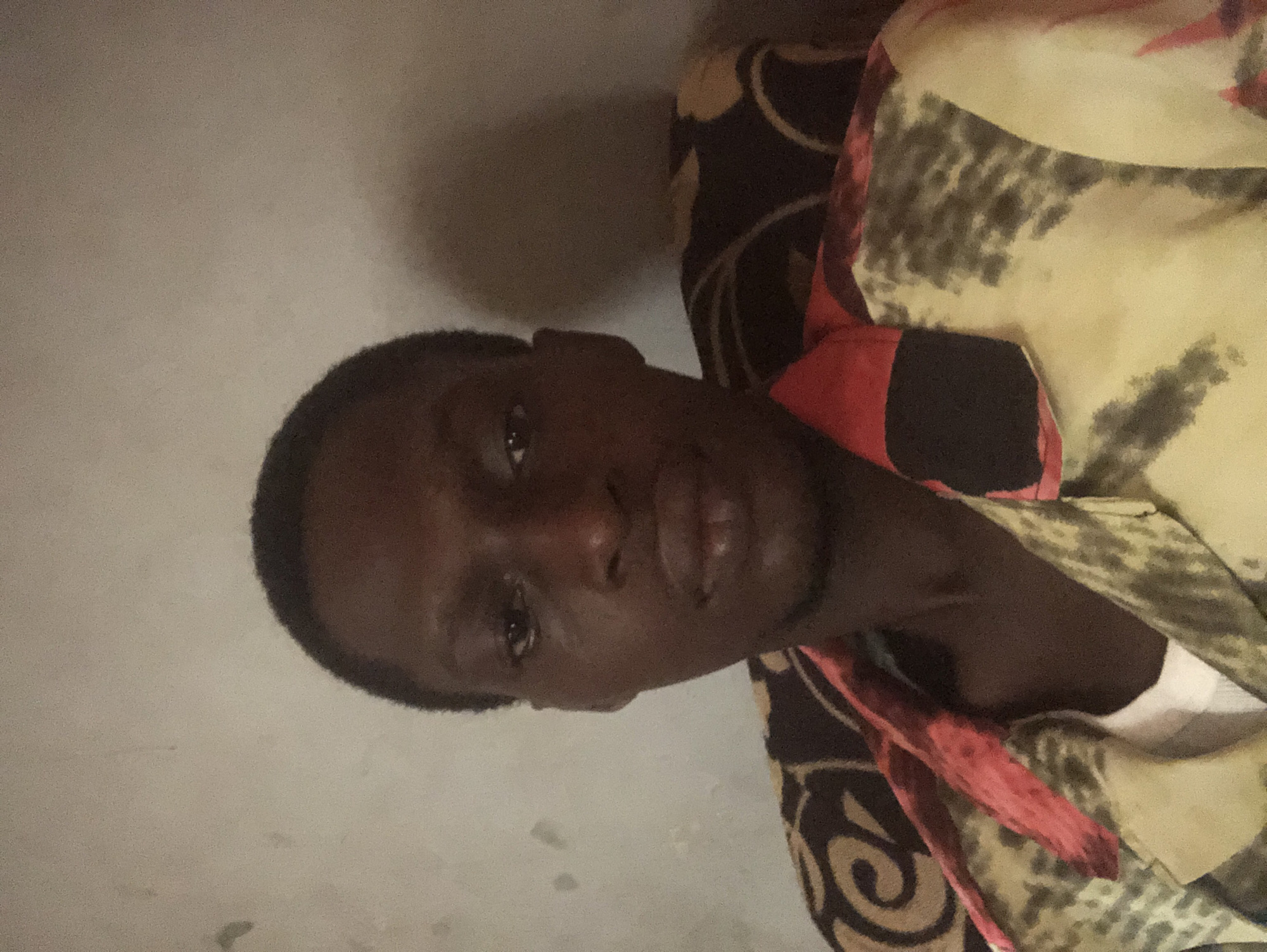 The image is a dark, slightly blurry, and grainy portrait of a person, originally captured at a 90-degree clockwise rotation. The subject is a young individual with dark skin, short dark hair, and a neatly trimmed beard or stubble. They are looking directly at the camera with a neutral to slightly serious expression. Their right eye, visible on the left side of the face from the viewer's perspective, shows a glint of light.
The person is wearing a patterned collared shirt. The fabric displays a complex design incorporating shades of beige, dark brown, red, and possibly black, with abstract shapes, swirls, and splotches. A piece of the collar and a red inner lining or trim are also visible.
The background consists of a plain, light-colored wall on the left side, which appears to be beige or light gray. On the right, behind the person's shoulder, a dark object with a similar intricate pattern, possibly a pillow or part of furniture, is visible. The dim lighting suggests an indoor setting, possibly during evening hours or in a poorly lit room. No specific activity is observed other than posing for the photograph. There is no visible text in the image.