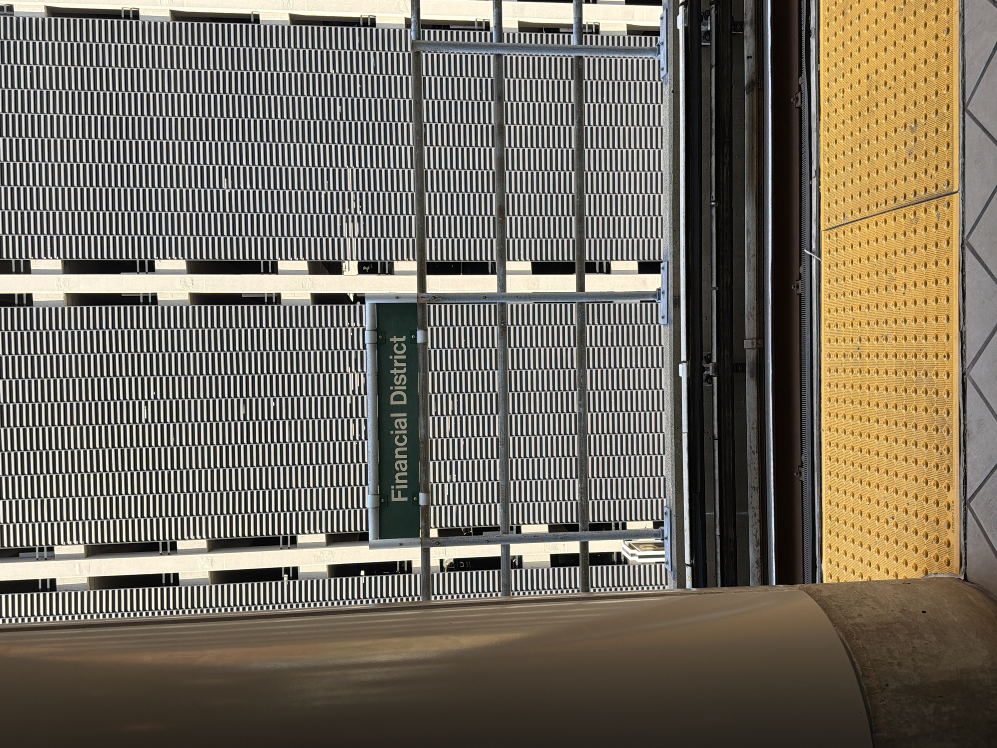 The image depicts a train station platform with a sign that reads "Financial District." In the foreground, a yellow textured tactile paving surface is visible, along with a section of train tracks. Behind the tracks, a metal railing separates the platform from the background. The background is dominated by a large building with a facade characterized by a repetitive pattern of horizontal dark and light stripes, suggesting a parking garage or a building with louvers. A thick, light-colored cylindrical pillar is positioned on the left side of the image, partially obscuring the building. The scene is captured in daylight, with strong shadows indicating a bright, sunny environment. The location is identified as being in Detroit, United States of America.