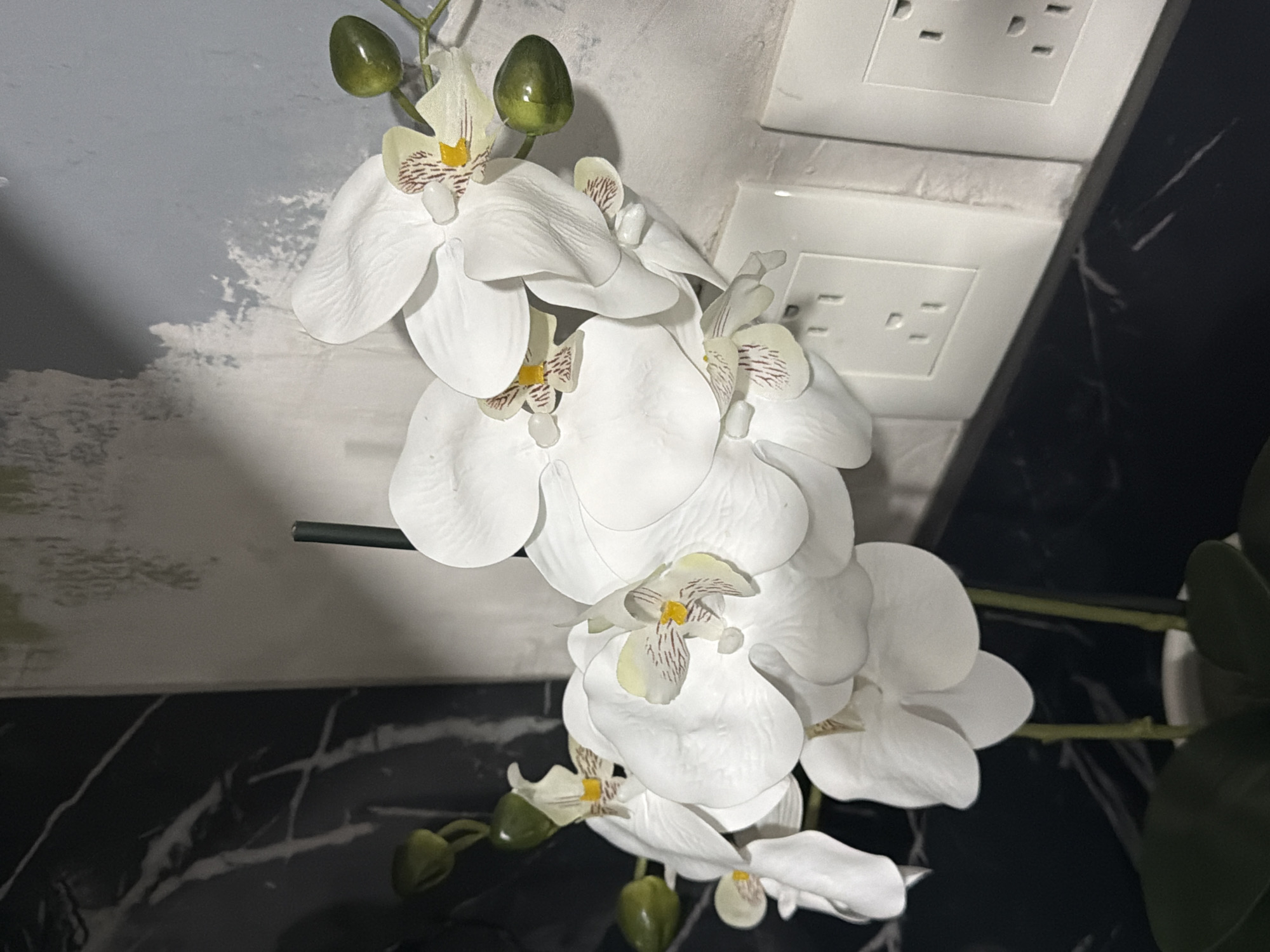 A close-up shot depicts a cascade of artificial white orchid blooms, punctuated by several unopened green buds. The background includes a textured, light-colored wall, adjacent to a pair of white electrical outlets. To the right, a dark, marbled surface is visible. The location and specific city are unknown.