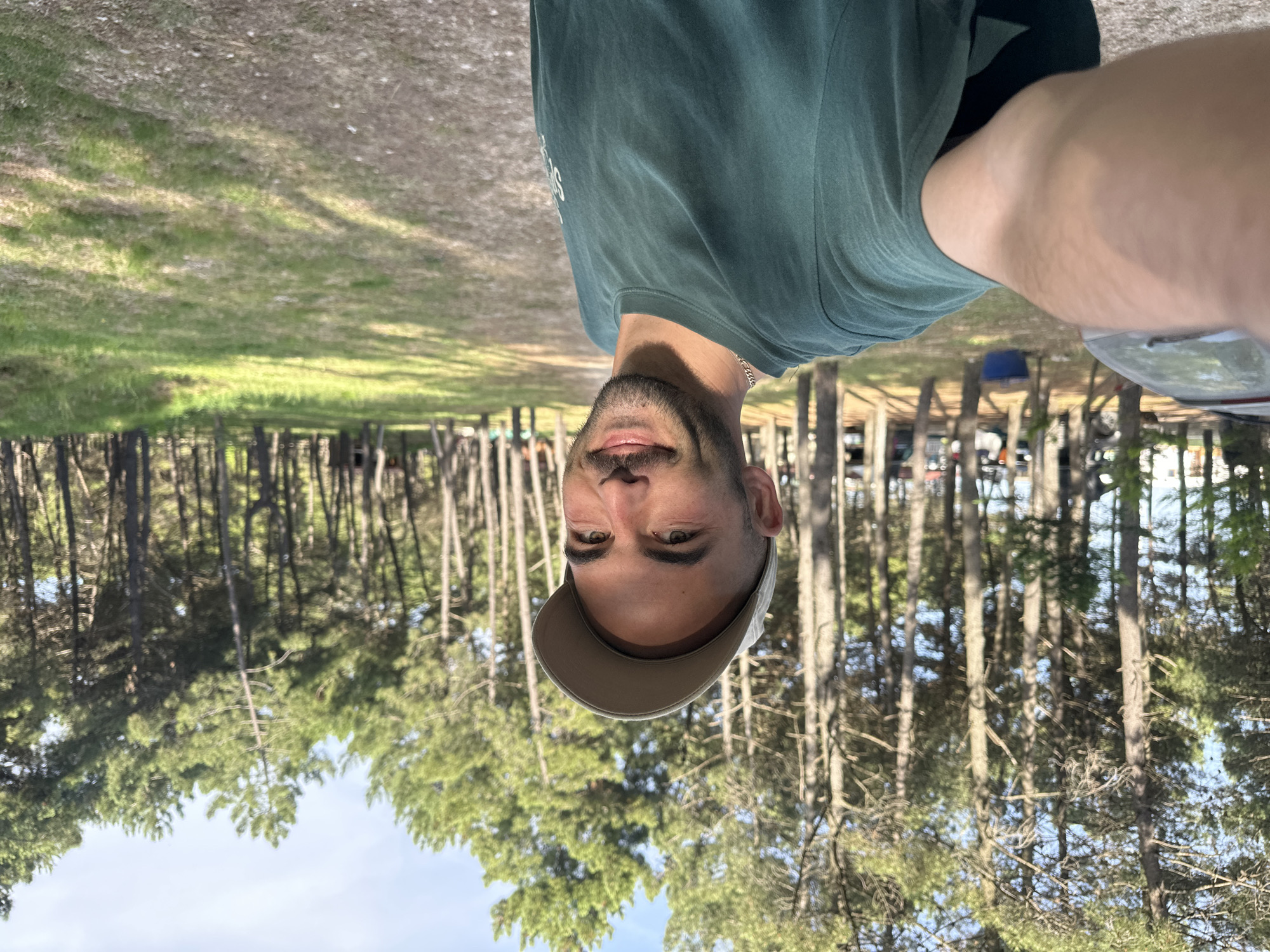 The image shows a man with a beard, wearing a green t-shirt and a tan baseball cap. He is facing the camera and appears to be taking a selfie. The background features a forest of tall, thin trees, possibly pine trees, with patches of grass and a light blue sky visible through the trees. The setting appears to be in a wooded area near Donji Štoj, Montenegro. In the background are vehicles and structures.