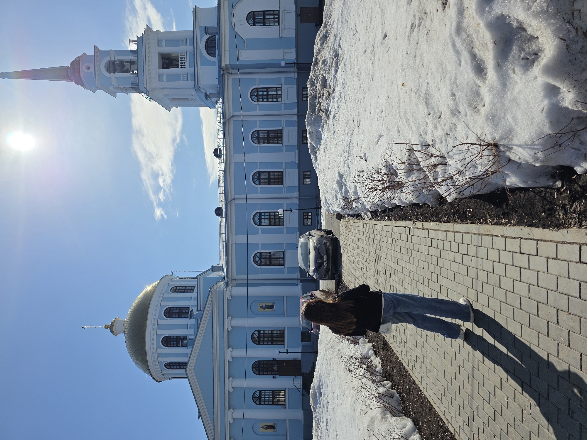 A daylight photograph depicts a scene in Votkinsk, Russia, featuring the Cathedral of the Annunciation (Blagoveshchensky Sobor) as the central subject. The prominent light blue and white religious edifice features a large, reflective golden dome topped with a cross and a tall, multi-tiered bell tower with a dark blue conical spire, golden finial, and visible bells. The building's facade is accented with white architectural trim, columns, arched windows with dark frames, and two framed religious icons.
The environment indicates late winter or early spring, with extensive patches of melting snow forming banks along a grey rectangular paver pathway. Exposed dark soil and bare deciduous shrubs are visible where snow has receded. The sky is clear blue with scattered wispy clouds, and the sun, positioned in the upper right, casts bright light and lens flare, creating a long shadow from the foreground subject.
A single individual is observed from behind, walking on the paved path towards the cathedral. The person has long, dark brown hair and is dressed in a dark jacket, a light-colored undershirt, blue jeans, and white sneakers. A white bag is carried over the left shoulder.
Two vehicles are parked near the building: a light grey Renault SUV with Russian license plate K771AX 18, and a partially obscured darker vehicle positioned to its left. Two dark street lamps are visible near the building's base.