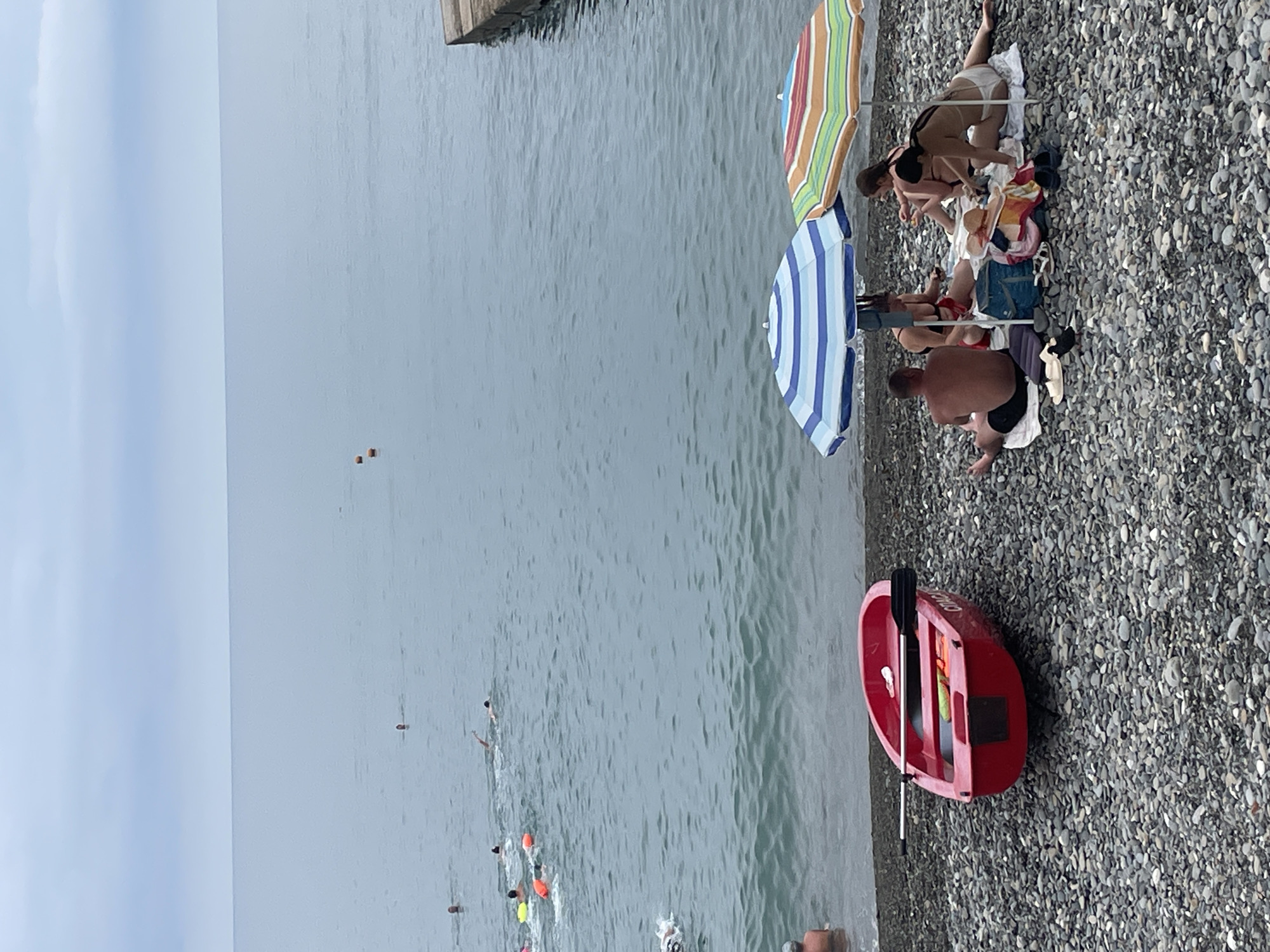 The image depicts a pebbled beach in Sochi, Russia, with the Black Sea in the background. A bright red rowboat with oars rests on the shore. Several people are visible in the water, swimming and appearing to be participating in some recreational activity, with a few buoys scattered in the distance. On the beach, a group of people are seated under two colorful beach umbrellas, one blue and white striped, and the other with multiple bright stripes. They appear to be relaxing, with beach towels and bags spread around them. The sky is overcast, contributing to a diffused light.