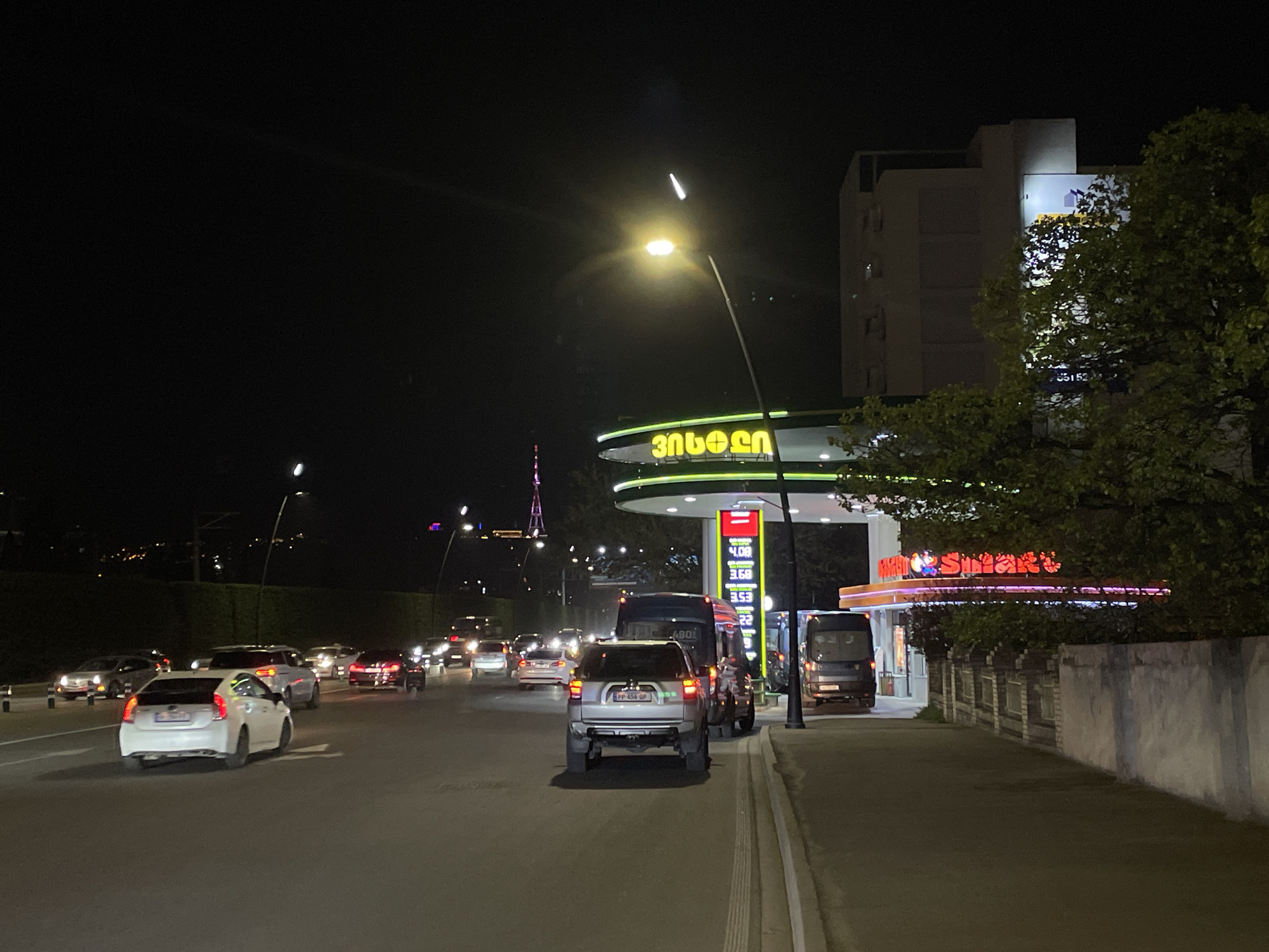 This nighttime image captures an active street scene in Tbilisi, Georgia, illuminated by artificial light sources. The primary subjects include a multi-lane road filled with vehicles, a gas station, and surrounding urban architecture and trees.
Numerous cars are visible on the road, with headlights and taillights creating streaks of light. In the foreground, a white Toyota Prius is in the left lane, while a silver SUV with the license plate "PP-454 OP" is positioned centrally, likely in traffic or approaching the gas station. Several other cars form a queue behind them. Two dark-colored vans or minibuses are also present near the gas station entrance.
A prominent gas station occupies the right side of the frame, featuring a distinctive curved canopy lit with bright yellow and green lights. The canopy displays text in Georgian script, which reads "ენერჯი" (Enerji), identifying the station brand. Visible on the fuel pump displays are prices such as "4.08," "3.68," and "3.53," likely denoting fuel costs in Georgian Lari. Adjacent to the pumps, a convenience store glows with a red neon sign that clearly spells out "Smart." Behind the gas station and partially obscured by trees, a multi-story building stands, with some illuminated windows and faintly visible numbers like "5152" on its facade.
The scene is clearly set at night, evidenced by the dark sky and the reliance on artificial lighting from streetlights, vehicle lights, and building signs. A bright streetlight directly illuminates the foreground, creating a lens flare effect. In the distant background on the left, a lit structure resembling a communication tower or monument, possibly the Tbilisi TV Tower, pierces the night sky. Green foliage from trees and hedges adds a natural element to the urban landscape. The image conveys a typical, bustling city night, with vehicles moving through a well-lit commercial area.
