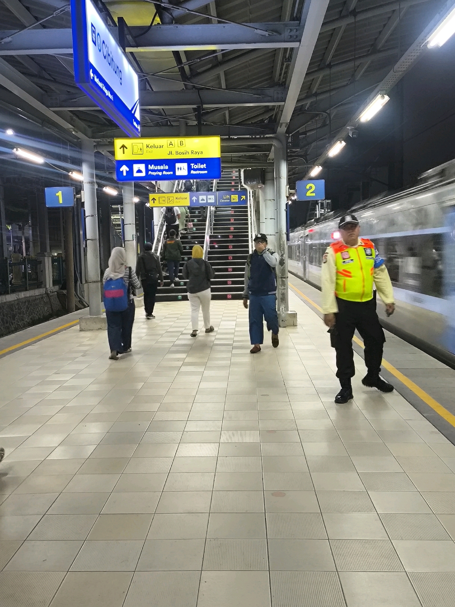 The image depicts a train station platform in Mekarsari, Indonesia. The platform is paved with light-colored square tiles and has a yellow line along the edge. A train is visible on the right, blurred due to motion. Several signs are present, including one that reads "Cibitung" in blue and white, indicating a destination. Other signs in yellow and blue provide directions to "Keluat" (Exit), "Musala" (Praying Room), and "Toilet". There are also signs for "Lift" and "Elevator".
In the foreground, a person wearing a yellow and green vest, dark pants, and a cap, possibly a railway employee or security guard, is walking towards the viewer. Behind them, several other individuals are walking on the platform. One person is wearing a hijab and carrying a backpack. Another individual is walking up a set of stairs. The overall environment suggests a busy public transportation hub. The lighting indicates it is either evening or indoors with artificial illumination.