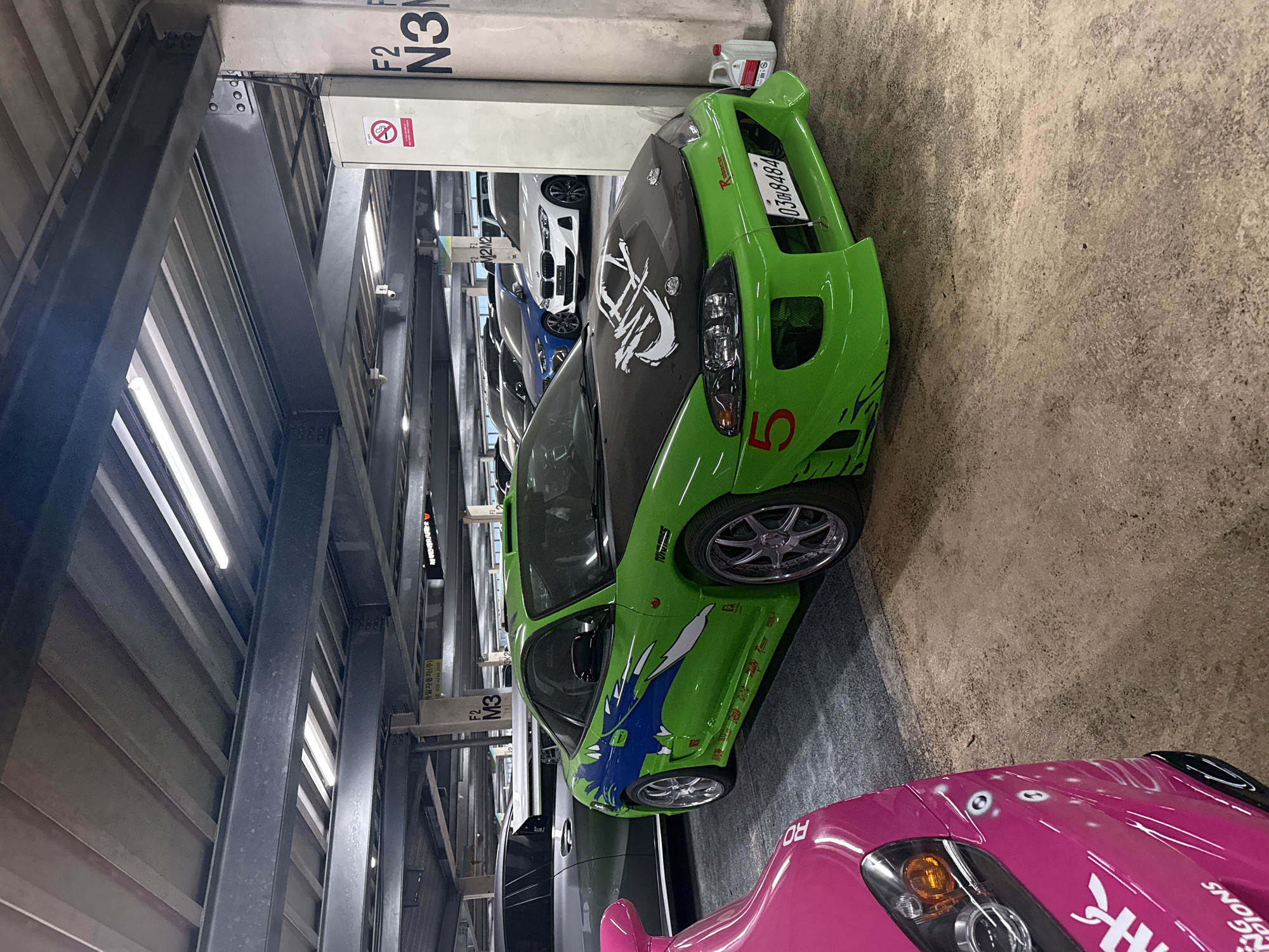 The image shows a green sports car with a black hood and a white design on the hood. The car has a license plate that reads "0308484". The number "5" is painted on the side of the car. The car is parked in a parking garage, with a pink car visible in the foreground. The parking garage is labeled "F2 N3" and "F2 M3". Text on one of the signs reads "사일자동차(주) 2010-4238-2040 100 M2M2". The location is presumed to be in Seoul, South Korea, based on the Korean text on the sign.