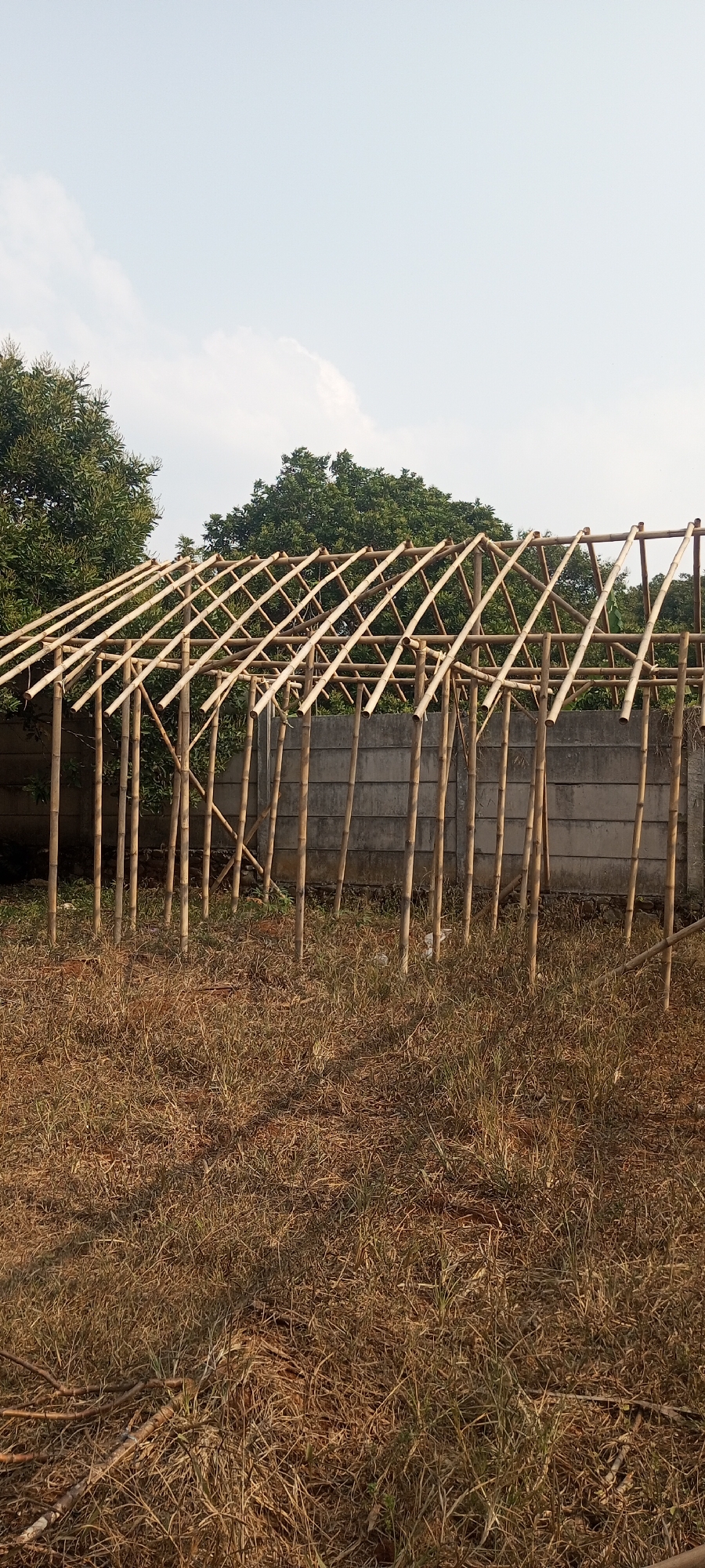 A partially constructed building frame made of bamboo poles and crossbeams stands in an outdoor, dry, grassy area in Taman Rahayu, Indonesia. The structure appears to be a lean-to or open shelter. A concrete wall runs horizontally behind the bamboo frame, and lush green trees are visible beyond the wall. The sky above is a pale blue with scattered white clouds. The ground is covered in dry, brown grass and fallen branches. No people are present in the image.