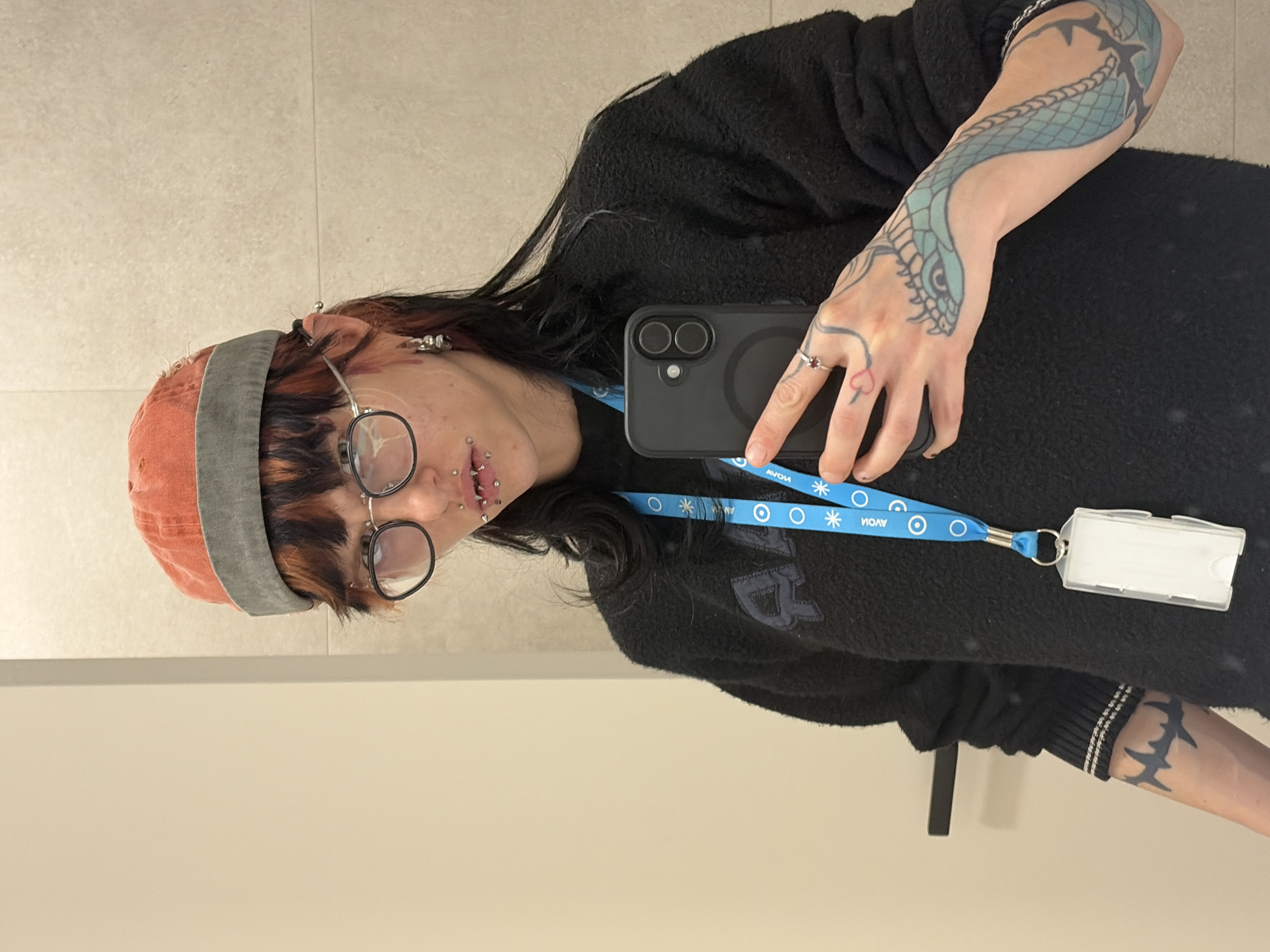 The image shows a person taking a mirror selfie indoors. The individual is looking directly into the camera, wearing black-rimmed glasses, a dark textured sweater, and an orange/red beanie with a grey band. Their dark hair has lighter, possibly dyed, auburn sections at the front.
Distinctive features include multiple facial piercings on the lips and ears, and several tattoos. A large blue and black snake design covers their right forearm, while a black barbed wire-like tattoo is visible on their lower left arm. A small red heart outline tattoo is on a finger of their left hand, which also bears a ring.
The person holds a black smartphone with a black case in their left hand, reflecting the scene. A blue lanyard, emblazoned with the text "AVON" and white circular and snowflake patterns, hangs around their neck, connected to a clear, empty ID holder.
The background is a tiled wall, featuring light beige or grey rectangular tiles, indicative of an interior setting such as a hallway or bathroom. There are no discernible cues for the time of day or specific weather. The overall impression is a casual, personal photograph.