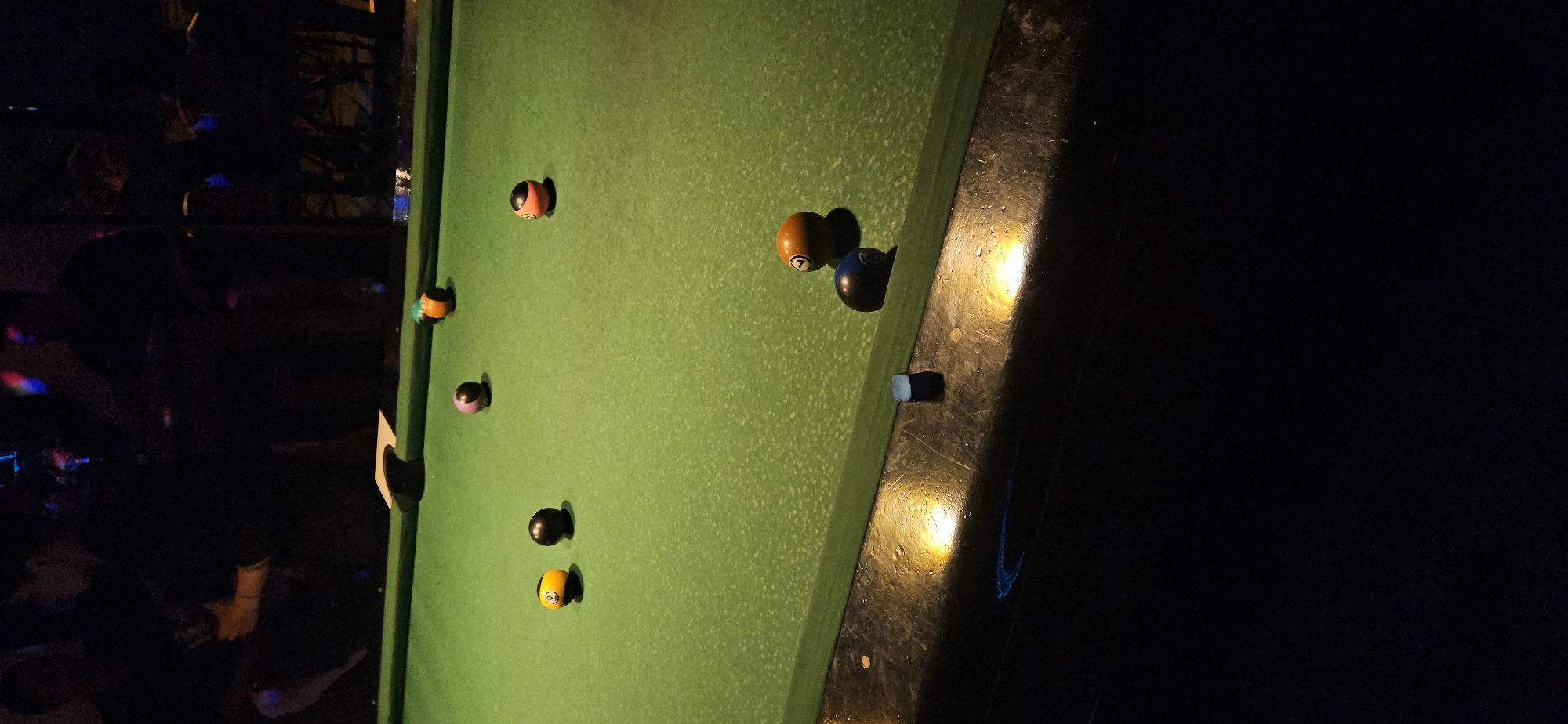 A low-angle, rotated shot depicts a pool table with scattered pool balls and a partially visible person in a dimly lit environment. The table has a green felt surface with multiple pool balls of varying colors, including orange, black, and purple, resting on it. A pool cue chalk cube is near the edge of the table. Adjacent to the pool table is a dark, reflective surface. In the background, a blurred figure of a person and vague outlines of furniture are visible. The lighting is uneven, with patches of light contrasting against deep shadows. The specific location in Nha Trang, Vietnam, is not identifiable from the image alone.