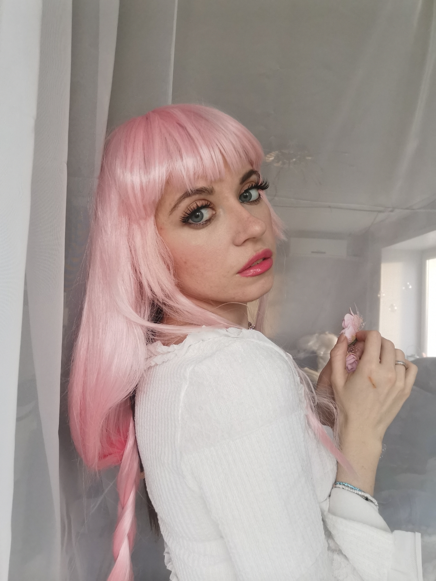 A young woman with striking pink hair, likely a wig, is captured in a medium close-up shot, looking directly over her right shoulder towards the camera. Her fair skin shows a scattering of freckles, and her eyes, a light blue-green, are enhanced with prominent false eyelashes and dark eyeliner. Her lips are painted a glossy hot pink. She is wearing a white, textured top with a ruffled neckline, possibly a sweater or blouse.
In her left hand, which is held delicately near her chest, she holds a cluster of soft pink artificial flowers or feathers, gently interacting with them with her fingers. A silver band ring is visible on her left ring finger, and two bracelets – one beaded in white and another thin blue/silver one – adorn her left wrist.
The background consists of soft, translucent white curtains on the left side of the frame, creating vertical folds. Behind her on the right, a light gray wall is visible, with an air conditioning unit mounted high on it. Further to the right, a window or another light source casts natural light into the room. The overall lighting is soft and even, suggesting an indoor setting during daylight hours. The atmosphere of the image is soft and somewhat dreamy, with the subject's gaze conveying a pensive or alluring expression. No visible text is present in the image.