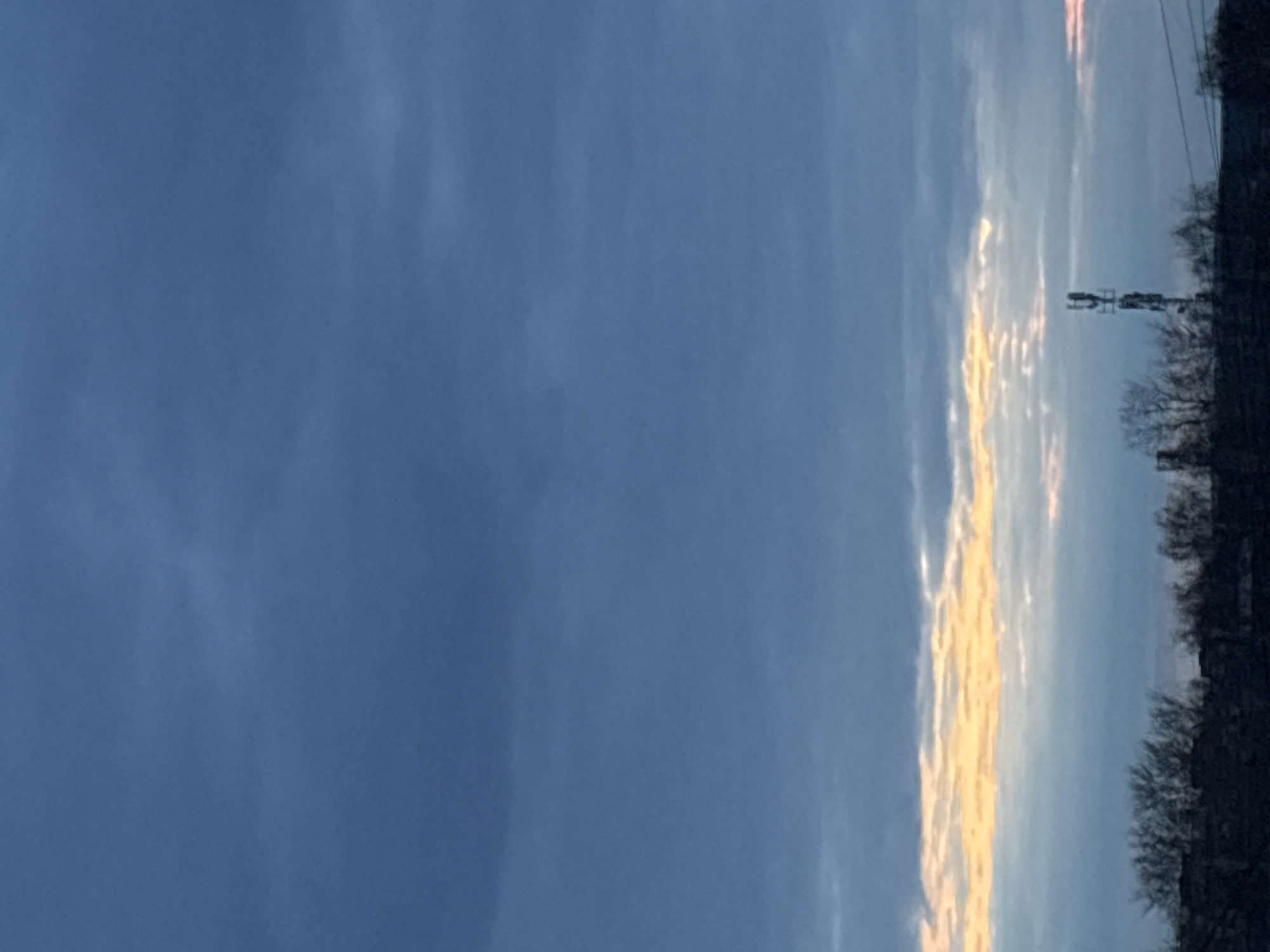 The image shows a landscape in Chaikovsky, Russia. A large portion of the image is occupied by a blue sky, with some clouds. On the right side, there are trees and some infrastructure, possibly power lines or a communication tower. Some clouds are illuminated with a golden light, indicating either sunrise or sunset.
