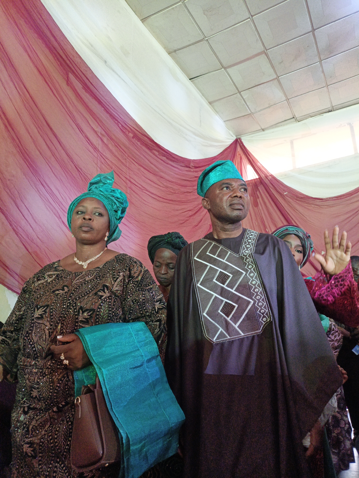 In Fagge D2, Nigeria, a medium shot captures a man and a woman standing indoors. The woman, wearing a brown patterned dress, a turquoise headwrap, and a diamond necklace, holds a turquoise shawl and a brown handbag. The man, beside her, wears a dark brown robe with geometric white embroidery on the chest and a matching turquoise hat. Behind them, draped maroon and white fabric serves as a backdrop. Other individuals are partially visible in the background. The setting suggests a special event or gathering.