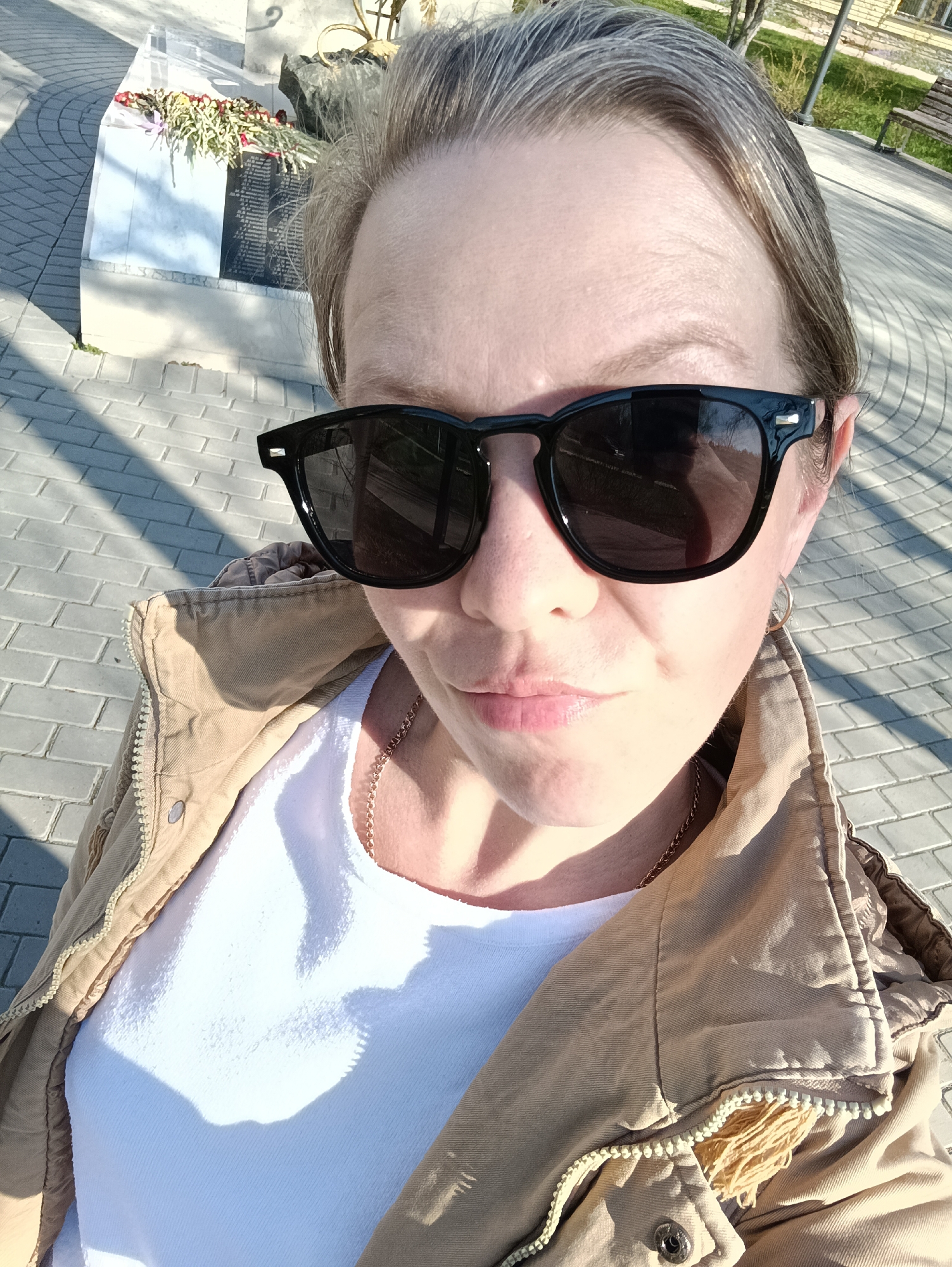 A person wearing sunglasses, a white t-shirt, and a tan jacket takes a selfie outdoors in Chaikovsky, Russia. The background includes paved ground, a monument with flowers, and a building. A gold necklace is visible around the person's neck. The day appears sunny.