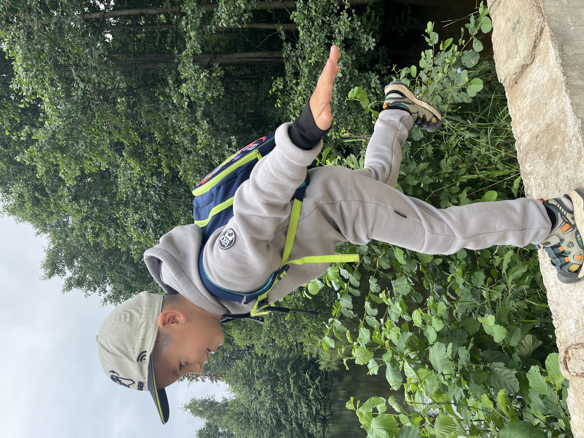 A young boy wearing a grey tracksuit, a blue and yellow backpack, and a grey baseball cap with a black brim stands balancing on a concrete ledge. His right arm is extended to his side, and his left leg is bent at the knee, with his foot lifted off the ground. He is smiling and looking to his left. Behind him is a body of water, likely a lake or river, and a dense forest with tall trees. The overall impression is of a child enjoying nature in an outdoor recreational setting. The scene is located in the city of Chaikovsky, Russia.