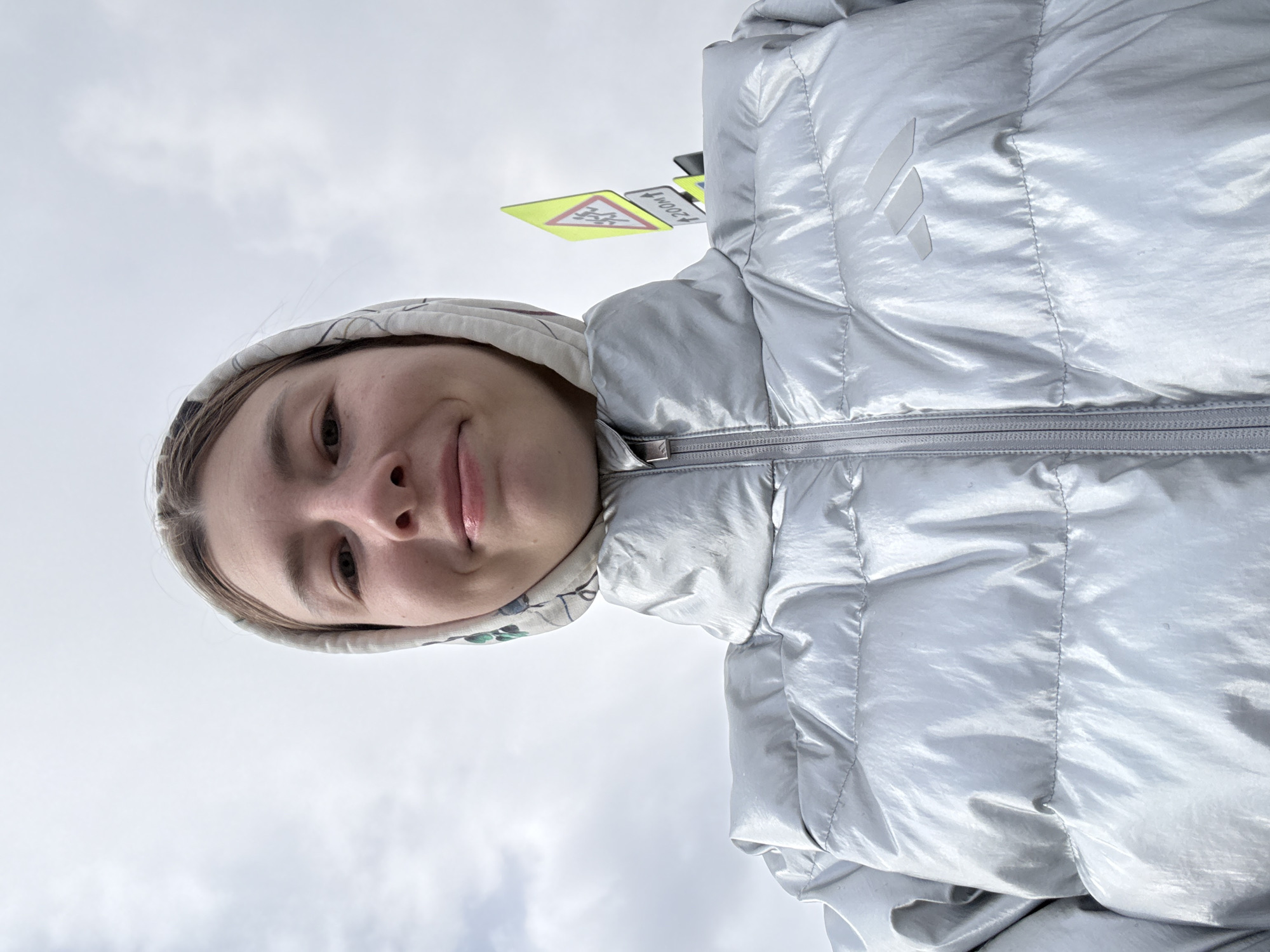 A young woman with fair skin is centered in the frame, her face displaying a slight smile. She is wearing a hooded jacket, silver in color, with an Adidas logo visible on the upper right chest. The hood is up, revealing a patterned fabric lining.
Behind and above her, slightly out of focus, are road signs written in the Cyrillic alphabet. The signs appear to be related to a school zone, with the text "200M" visible on one sign. The sky above is overcast.
The location is difficult to confirm definitively, but the road signs suggest a possible location in a Russian city. Given the request, Moscow is a plausible guess, however, there is not enough detail to know for sure.