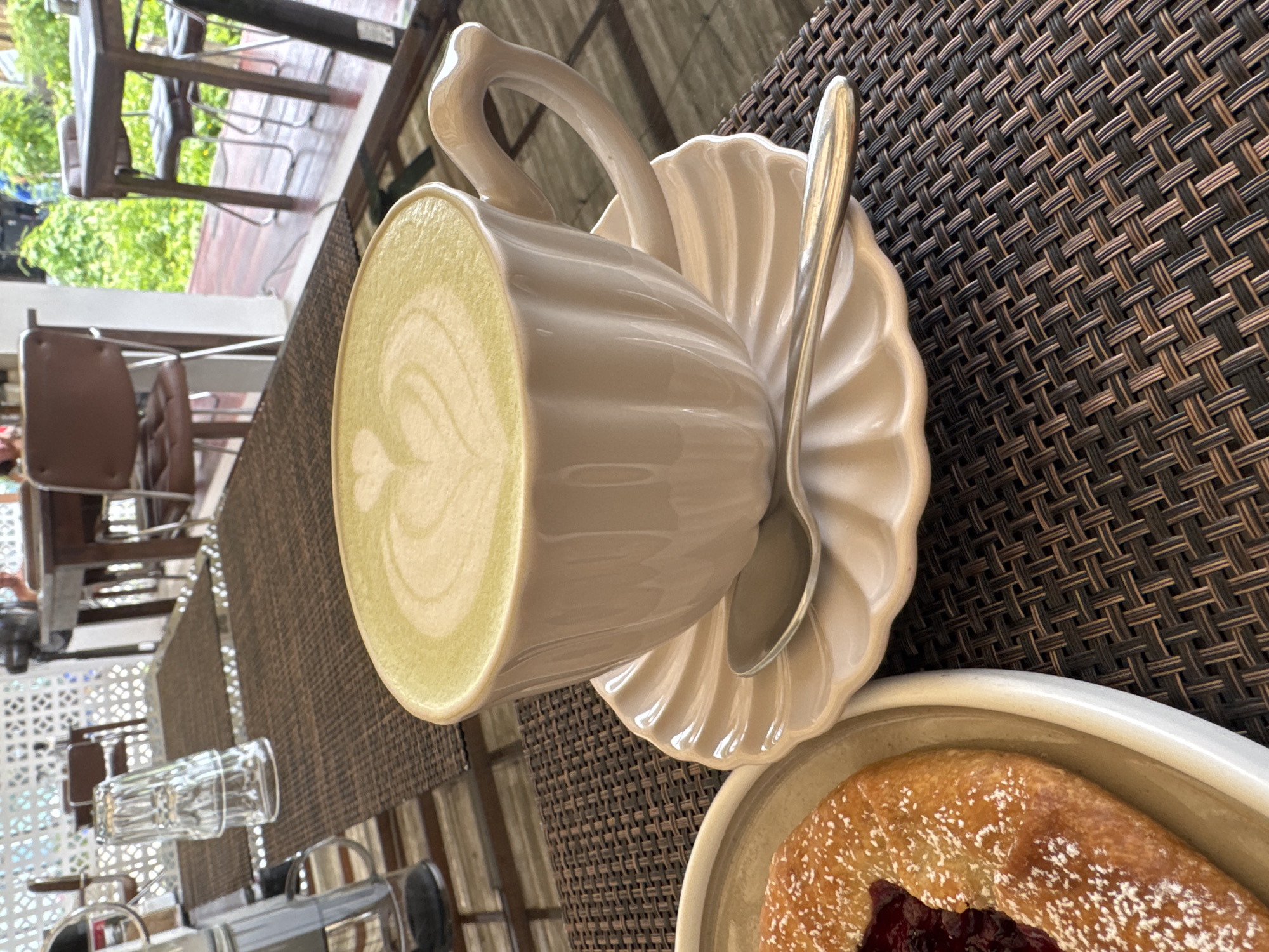 In Baan Mae Nam, Thailand, a close-up shot focuses on a cup of matcha tea with latte art and a pastry on a table. The matcha is a vibrant green with a heart-shaped design on top, served in a white ceramic cup with a fluted saucer. A silver spoon rests on the saucer. To the left, a portion of a golden-brown pastry, dusted with powdered sugar and filled with a red jam, is visible on a white plate. The table is covered with a dark brown woven placemat. In the background, blurred glimpses of outdoor seating and lush greenery suggest a cafe or restaurant setting, with tables and chairs visible.