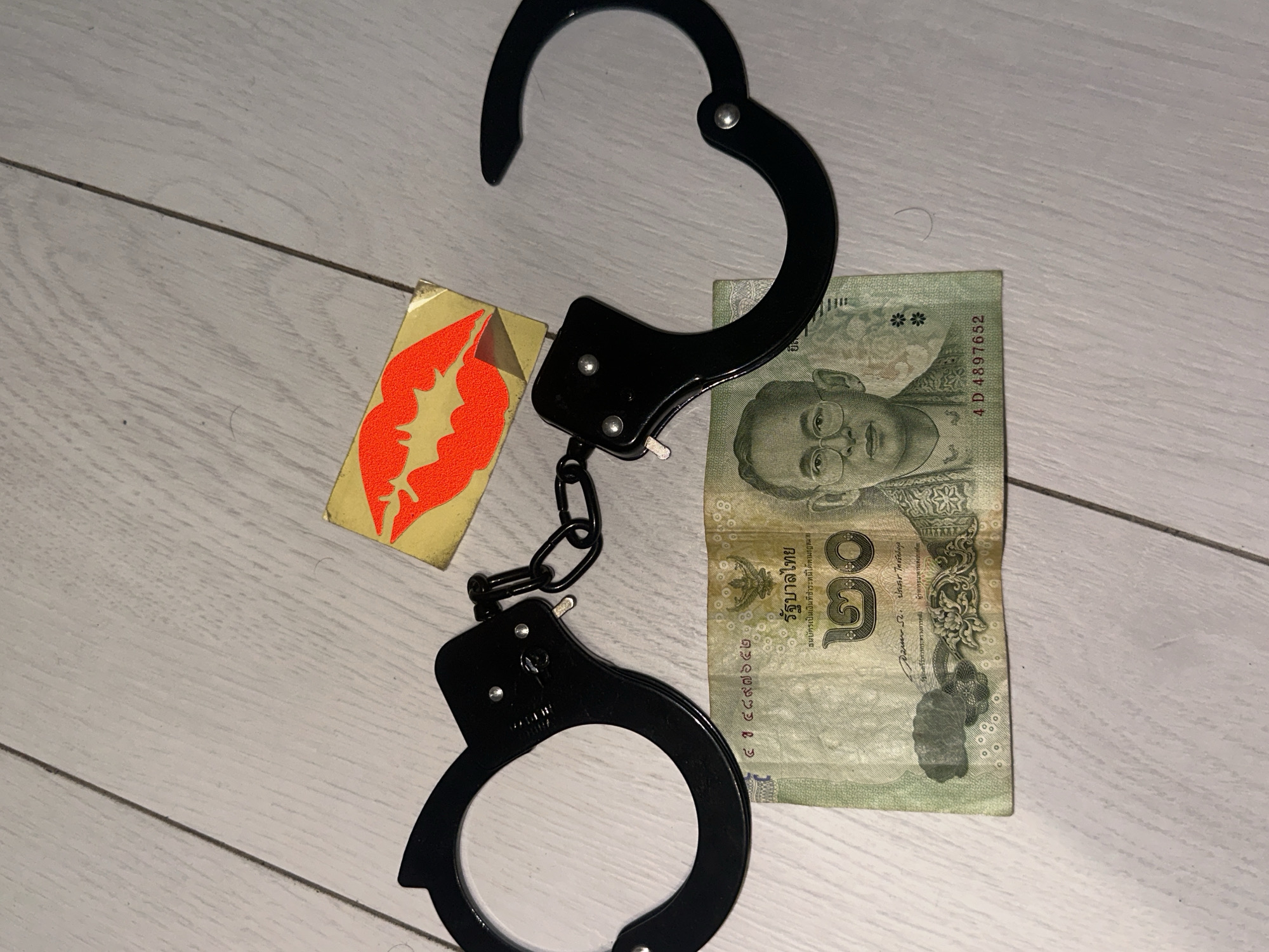 A close-up, overhead shot shows a pair of black metal handcuffs, a twenty baht Thai banknote, and a sticker of red lips on a light-colored, wood-planked floor. The handcuffs are open and appear to be lying flat on the floor, with one cuff to the left and the other to the right, connected by a chain. The banknote is positioned horizontally between the cuffs. The red lips sticker, with a yellow background, is placed to the top right of the handcuffs. The scene is well-lit, with shadows cast by the objects. The background consists of evenly spaced, light-colored wooden planks with visible grain and a subtle sheen. There is no indication of the location in Sofia, Bulgaria within the image itself.