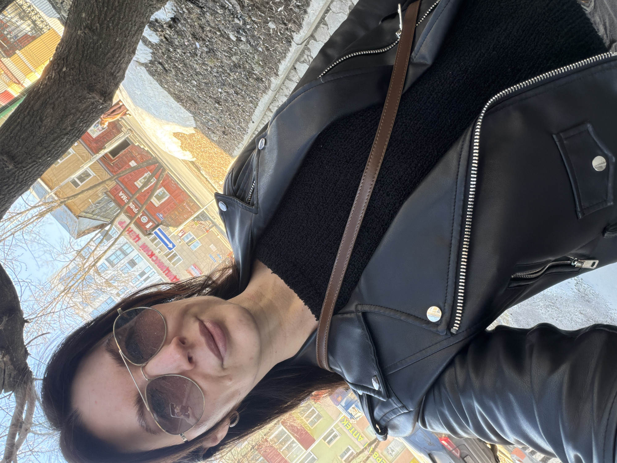 A woman with sunglasses is outdoors, possibly on a sidewalk, wearing a black leather jacket over a black textured top, with a brown shoulder bag strap visible. The background shows buildings, possibly in Perm, Russia, based on Cyrillic script signage on the buildings. Bare trees are in the background suggesting it is winter or early spring.