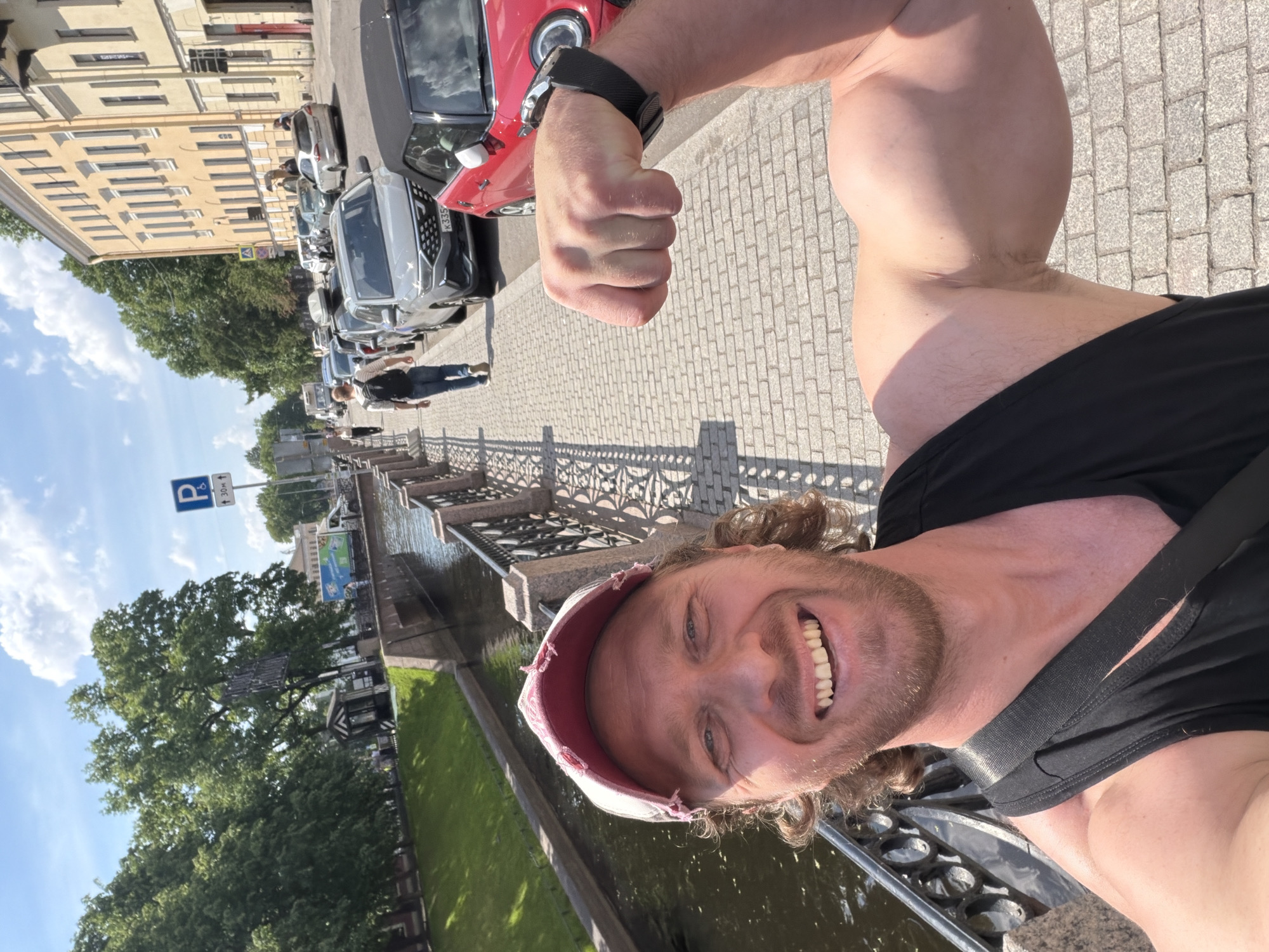 This selfie captures a cheerful man flexing his bicep on a sunny day in Saint Petersburg, Russia. The man, with curly light brown hair and a beard, smiles widely at the camera, wearing a distressed light-colored baseball cap, a black tank top, and dark athletic pants. A smartwatch is visible on his left wrist.
The scene unfolds along a canal, with an ornate metal railing separating the waterway from a cobblestone sidewalk. Behind the railing, large green trees line the canal bank. Across the canal, a street with several parked cars is visible, including a red Mini Cooper with a license plate partially reading "K335". Further down the street stands a multi-story yellow building featuring numerous rectangular windows. A blue "P" parking sign with "↑ 30m ↓" indicates parking is allowed for 30 meters in both directions. The sky is bright blue with scattered white clouds, suggesting clear, fair weather and strong daylight, evidenced by crisp shadows. The man's happy expression conveys a sense of enjoyment and confidence.