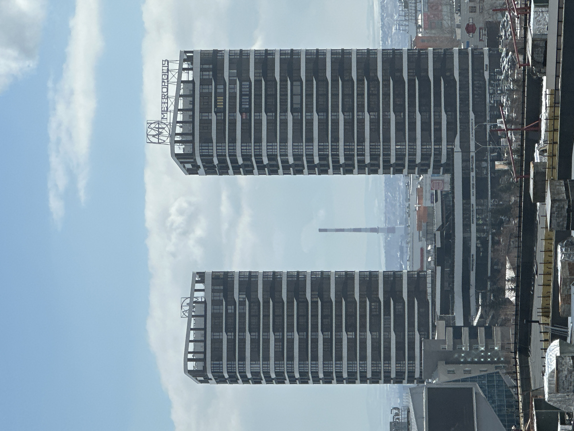 The image, when viewed in its correct landscape orientation, presents a modern urban landscape dominated by two tall, sleek high-rise buildings under a bright, partly cloudy sky.
The most prominent feature is the taller building on the left, adorned with a large rooftop sign. The sign clearly displays a stylized "M" logo followed by the text "METROPOLIS". This building, and a slightly shorter one to its right, exhibit a contemporary architectural design with repetitive patterns of dark windows and light-colored vertical facade elements, suggesting balconies or structural divisions.
The background reveals a broader cityscape, featuring numerous other buildings, some appearing residential and others commercial. Elements of ongoing construction, including red scaffolding or cranes, are visible in the middle-right and far-right portions of the image, indicating development in the area. A distant industrial smokestack rises against the horizon.
The sky is a clear blue, streaked with elongated white clouds, suggesting a daytime scene with good visibility. No people or specific human activities are discernibly taking place, placing the focus on the urban infrastructure. A faint red hexagonal "STOP" sign can be observed on a building in the mid-right background. The setting is Krasnoyarsk, Russia.