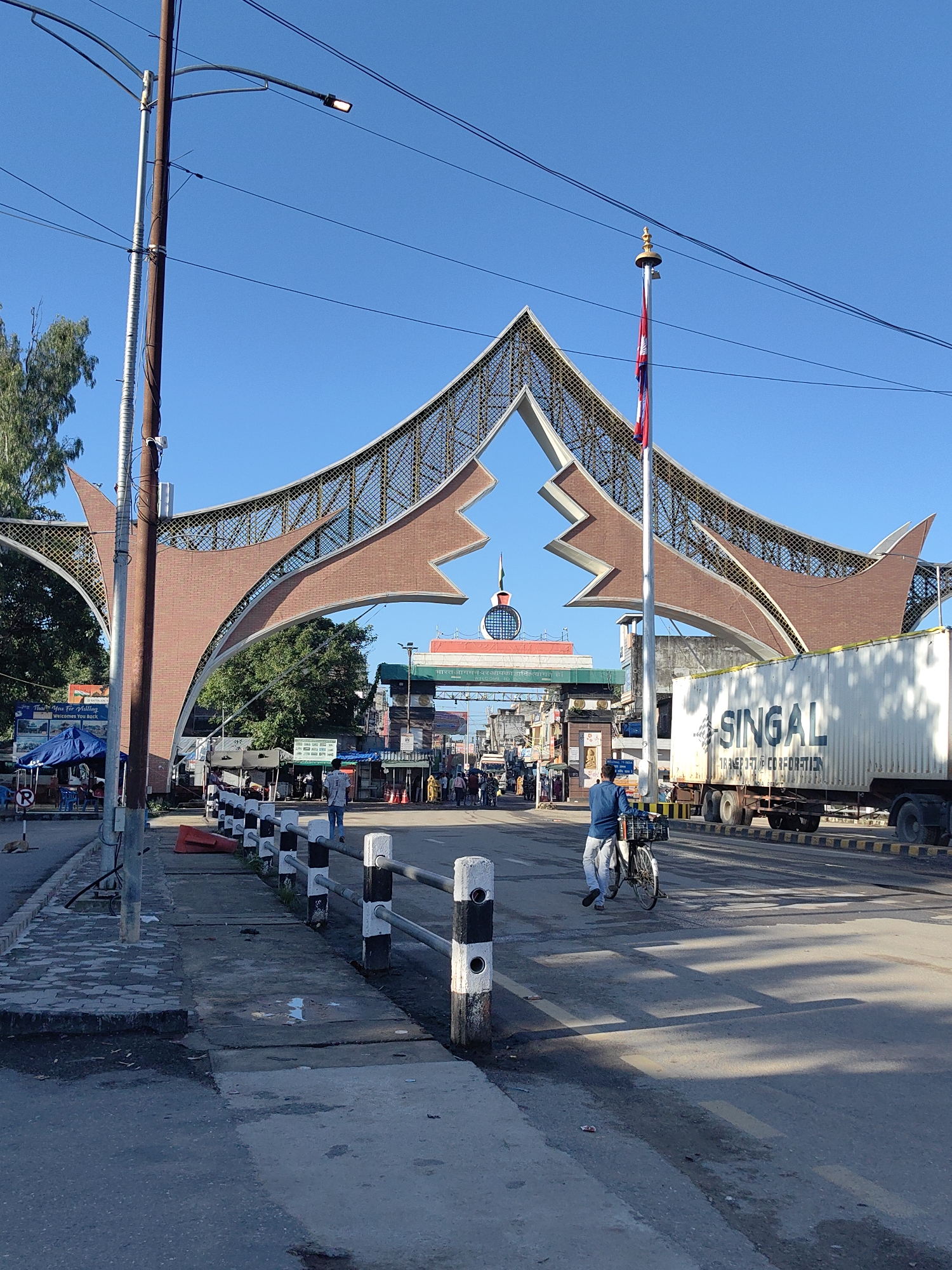 The image depicts the Belahiya border crossing in Belahiya, Nepal, under a clear blue sky. A large, modern archway with brick and metal detailing spans the road. A Nepali flag flies from a tall flagpole to the right of the arch. In the foreground, a man on a bicycle, laden with a basket on the back, cycles away from the camera. To the right, a large white shipping container truck with "SINGAL TRANSPORT CORPORATION" written on its side is parked. Several small stalls and shops are visible under the archway, with people milling about. A black and white striped barrier runs along the side of the road. The overall scene suggests a busy border point with ongoing commercial activity.