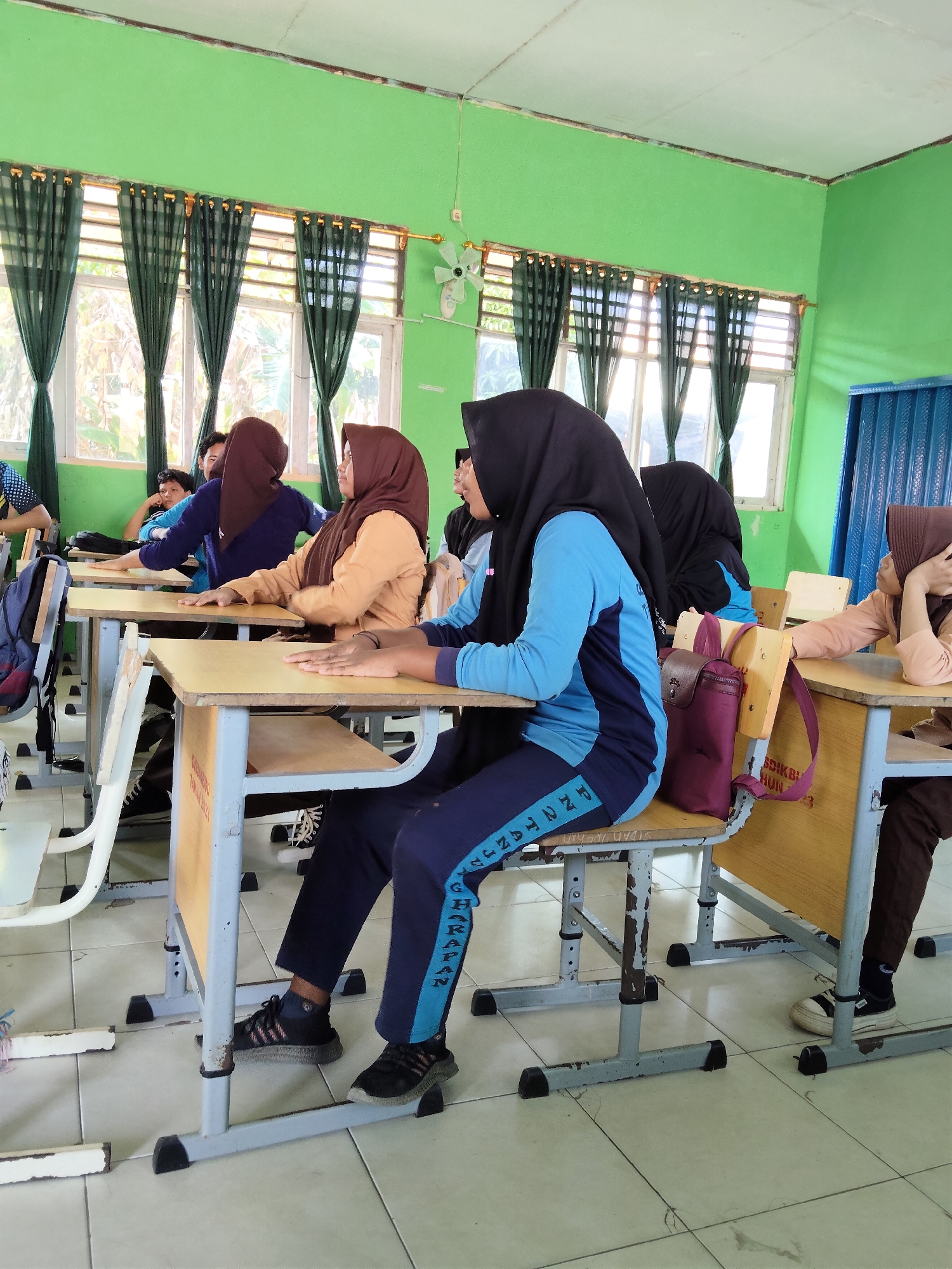 The image shows a classroom in a school, possibly in Lori, Indonesia, with several young female students wearing hijabs and school uniforms. The classroom has green walls and windows with dark green curtains. Desks and chairs are arranged in rows. One student in the foreground is wearing a blue and navy blue uniform and a black hijab. She is sitting at a desk, looking to the right. Another student next to her is wearing a brown hijab and a light brown uniform, also seated at a desk. Other students are visible in the background, some looking forward, others turned away. The floor is tiled. A fan is mounted on the wall near the windows. Bags are placed on some of the desks and chairs. The overall impression is of a typical school classroom scene.