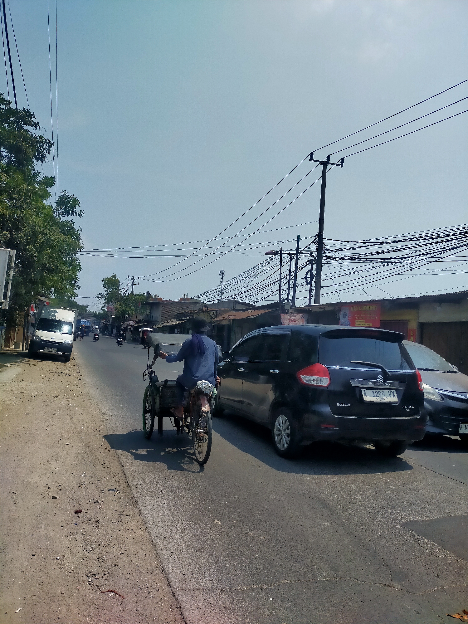 The image captures a street scene on a sunny day with a clear sky. In the foreground, a person is riding a three-wheeled vehicle, possibly a pedicab or a modified bicycle with a cargo area. The vehicle has two wheels at the back and one at the front, and the rider is wearing a dark blue shirt and a hat. The vehicle is on the right side of the road, moving away from the viewer.
The road is paved and appears to be a two-lane road with traffic. Several cars are visible on the road. A black Suzuki Ertiga is in the right lane, with its license plate reading "A 1299 VX". To the right of it, a portion of another car is visible. In the left lane, a white van is parked on the side of the road.
The left side of the road features a dirt shoulder with scattered debris. Trees with green foliage are growing along the side of the road. In the background, there are buildings with corrugated metal roofs and various signs, including one that reads "LAUNDRY". Numerous power lines and utility poles are prominent in the background, suggesting an urban or suburban environment. The overall impression is a typical street scene in a moderately busy area.