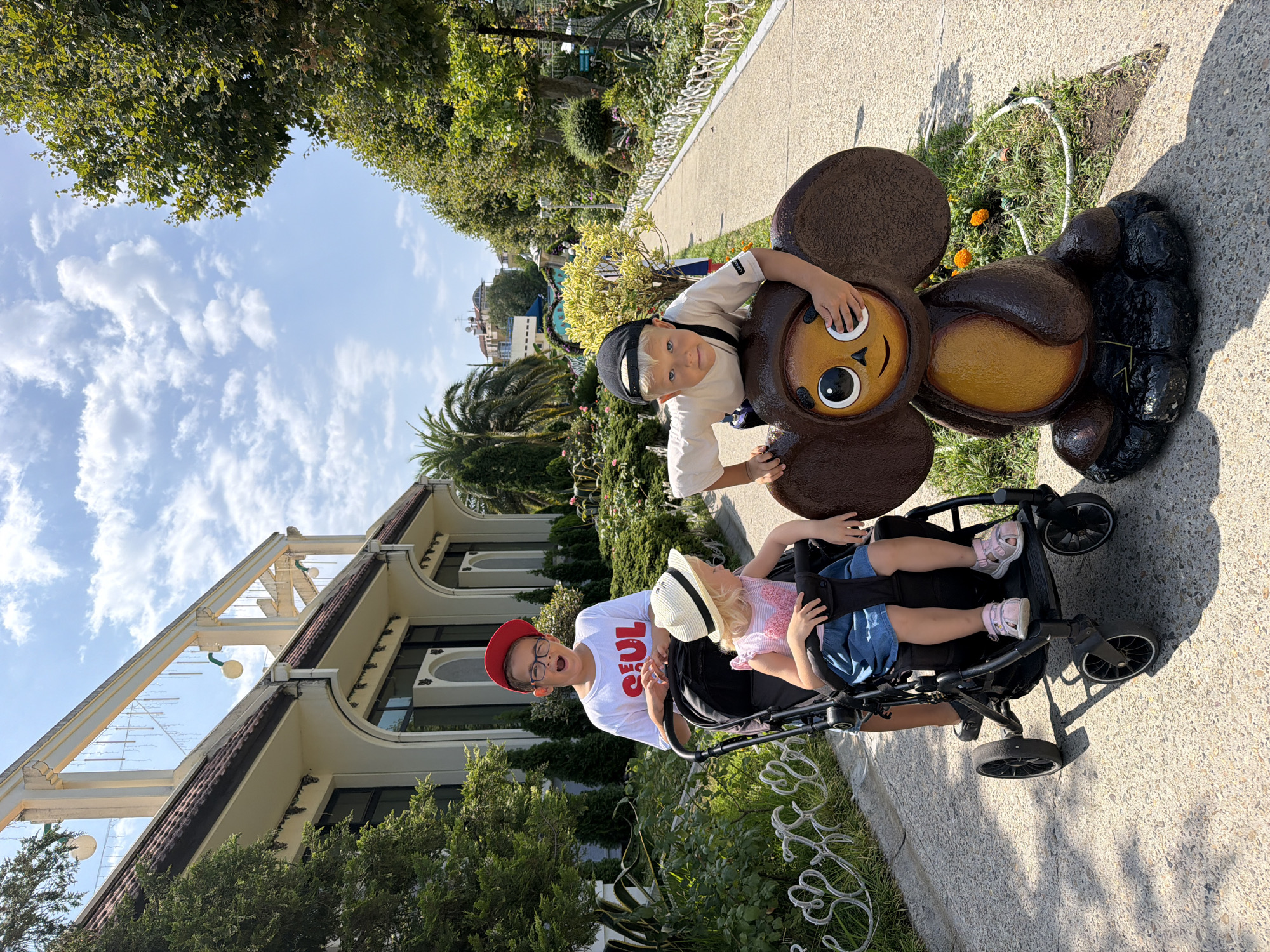 In an outdoor park setting in Sochi, Russia, a boy wearing a red baseball cap and glasses poses with his mouth open, leaning on a stroller occupied by a young girl in a white hat. The girl is wearing a pink top and a denim skirt. Behind them, another boy wearing a black baseball cap and a light-colored t-shirt leans on a large, brown, cartoonish statue of a character with big ears, covering one eye. The statue is positioned on a grassy area with small orange flowers. The park features a paved walkway, lush greenery, trees, and a building with white columns and arched windows in the background. The sky is blue with scattered clouds.