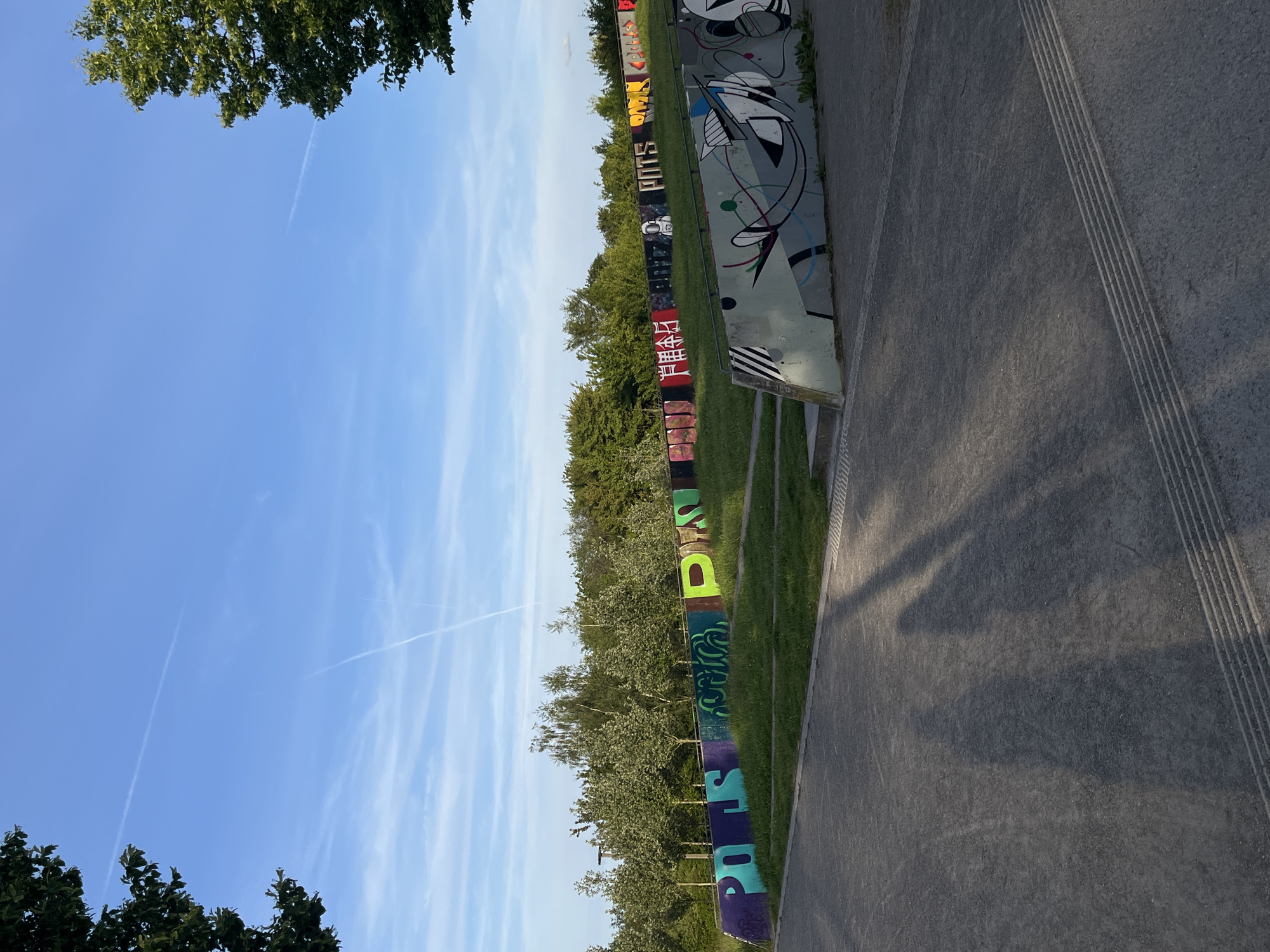 The image shows a concrete pathway running along the edge of a grassy area in Landau in der Pfalz, Germany. Along the grassy area is a low wall, which is covered with graffiti art in various styles and colors. Beyond the wall, there are trees and a blue sky with some white clouds. The pathway has a textured surface with parallel lines, and shadows cast by trees are visible on the concrete.