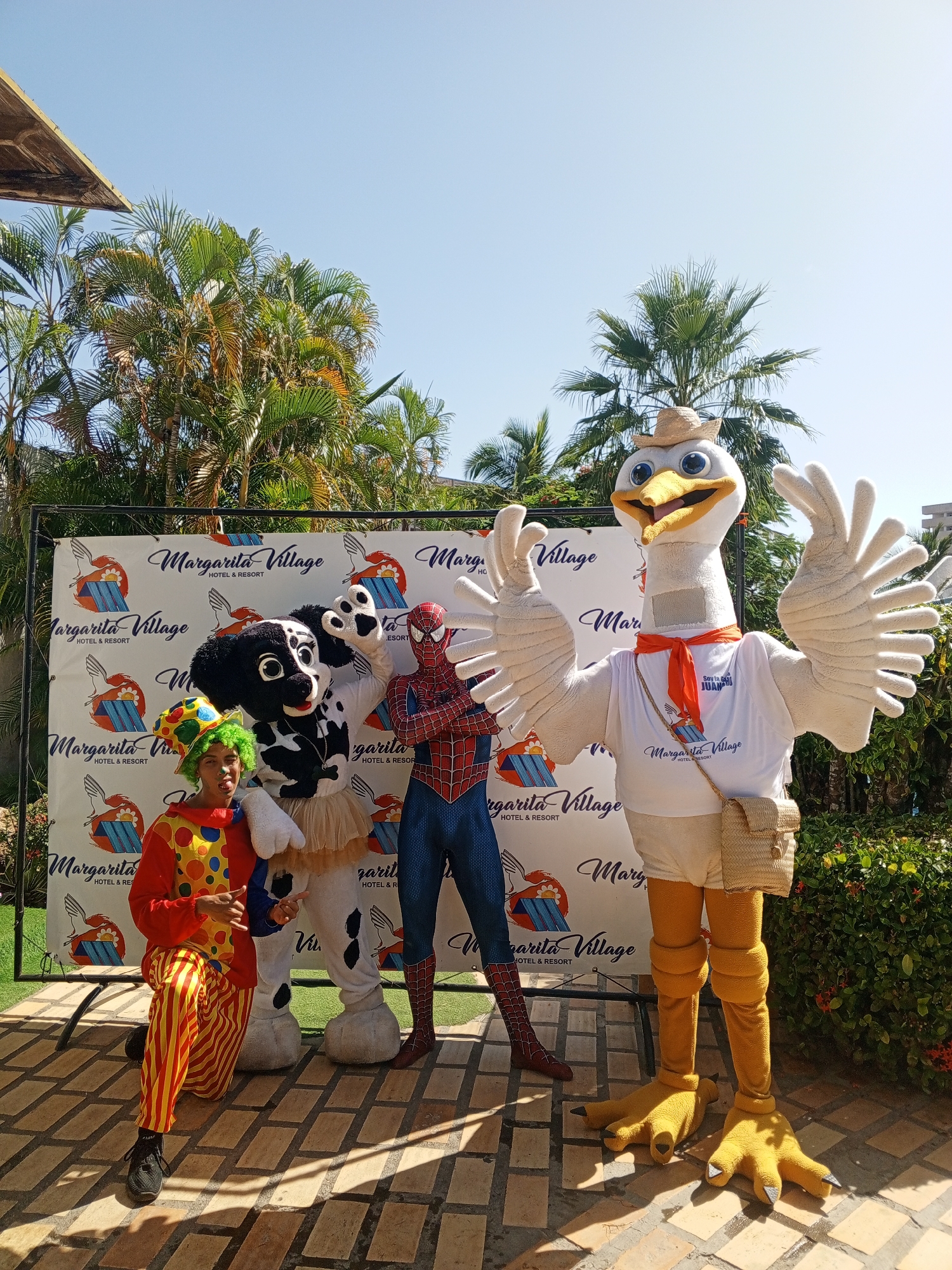 The image shows a group of costumed characters posing in front of a "Margarita Village Hotel & Resort" branded backdrop. The characters include a clown in a red and yellow striped suit and a green wig, a dog character, Spiderman, and a large bird mascot wearing a straw hat, a "Margarita Village" t-shirt, and a satchel. The backdrop has the logo of Margarita Village Hotel & Resort repeated across it. The setting appears to be an outdoor patio area with a brick-patterned ground, palm trees and other greenery in the background, with the blue sky visible above. The sign in the background states "Margarita Village Hotel & Resort" with one of the characters wearing a shirt that says "Soy de San Juan," indicating that the location may be in or near San Juan Bautista on Margarita Island, Venezuela. Considering this island location and the branding behind the characters, it is reasonable to assume the photograph was taken at the Margarita Village Hotel & Resort near the city of Porlamar, Venezuela.