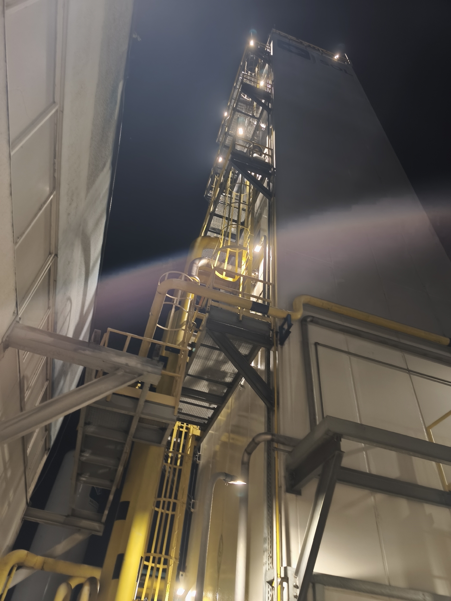 A nighttime, low-angle shot captures the exterior of an industrial facility. A tall, light-colored structure dominates the right side of the frame, appearing to be a storage tank or silo, with a dark, abstract logo on its upper portion. A complex network of yellow pipes and a metal external staircase with yellow railings ascend the side of this structure. The staircase features gridded metal flooring and is illuminated by several bright overhead lights. To the left, another building with a textured, light-colored facade is visible, running parallel to the main structure. This building also has visible structural elements and pipes. The overall scene suggests an operational industrial area at night, with artificial lighting highlighting the machinery and infrastructure. The sky is dark and appears clear.