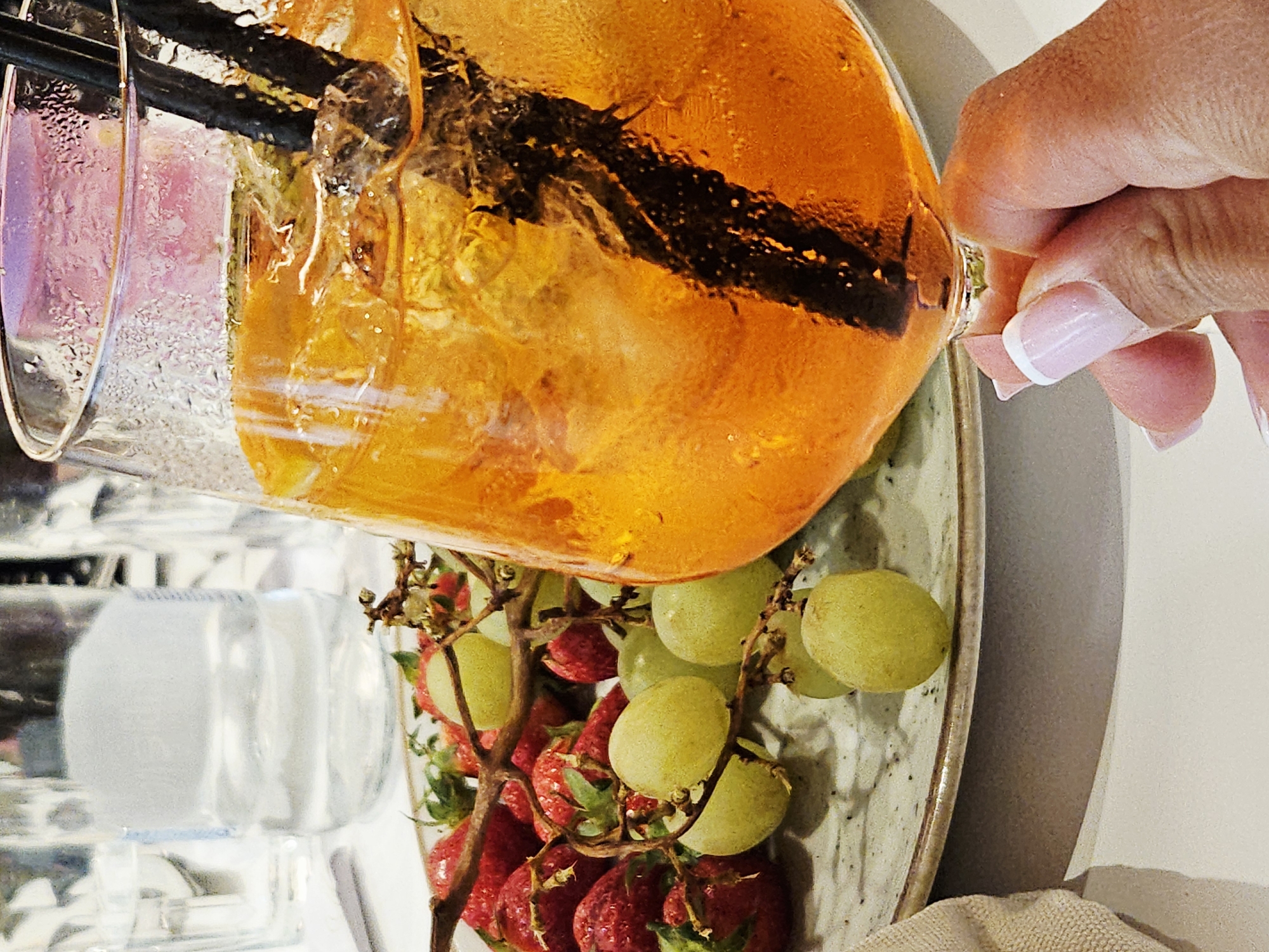 A tilted eye-level shot presents a refreshing beverage and fruit arrangement. A hand with manicured nails holds the rim of a large glass filled with an orange liquid and straws, positioned at the top right of the frame. Below the glass, on a plate, is a medley of strawberries and grapes. In the background, a few additional glasses are visible, suggesting a bar or dining setting. The photograph suggests an upscale dining experience, potentially at an establishment in Unknown, Unknown.