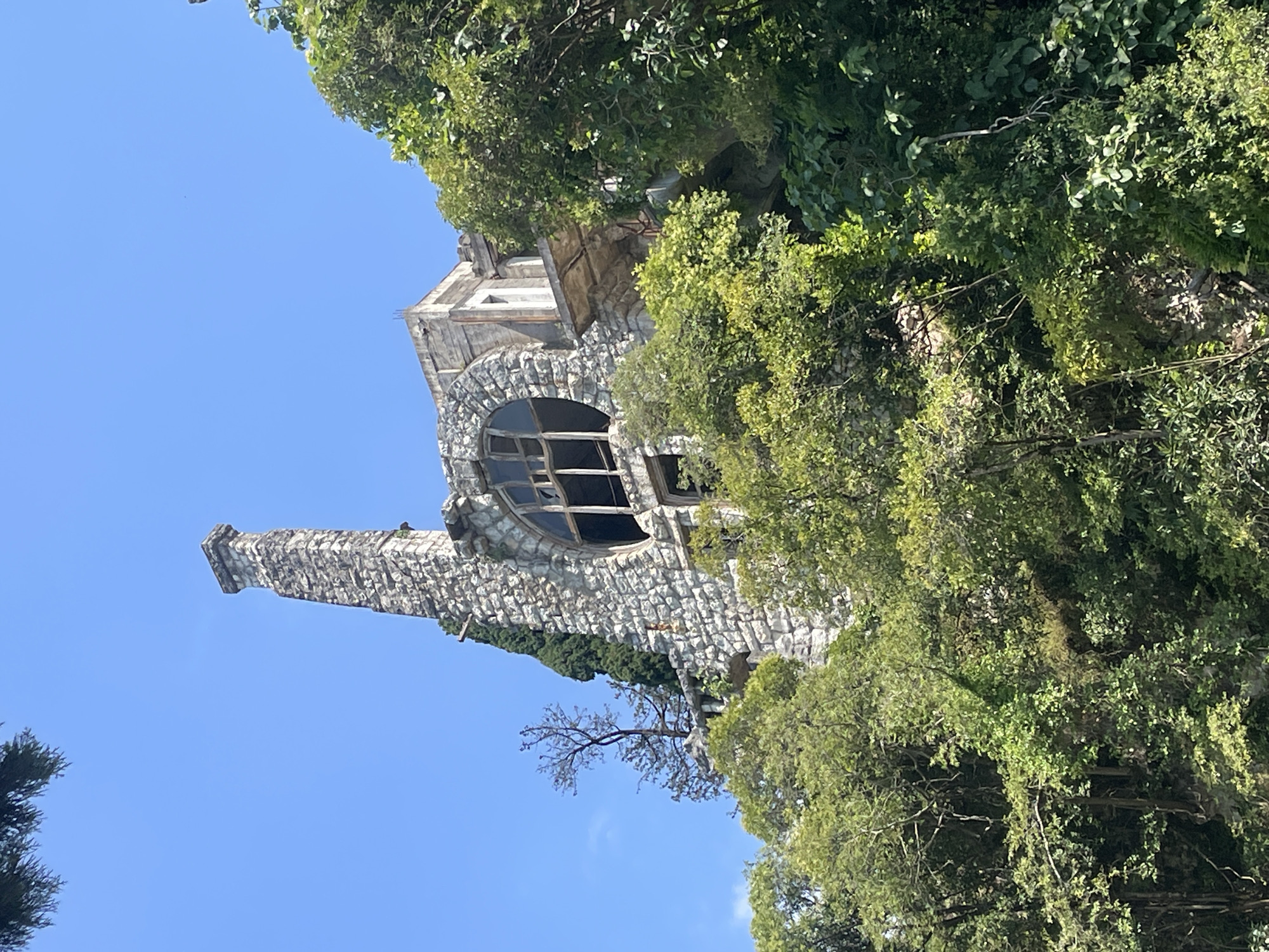 The image shows the abandoned and partially collapsed D.I. Gvazava Mansion, an architectural landmark located in Abaata, Abkhazia. The mansion is situated on a hillside amidst dense foliage. The structure is made of rough-hewn stone and features a prominent, tall chimney. A large, circular window with broken panes is visible on the facade. A balcony extends outwards, also appearing deteriorated. The surrounding environment is dominated by lush green trees and vegetation, with a clear blue sky overhead. There are no people visible in the image. The scene conveys a sense of decay and disuse.