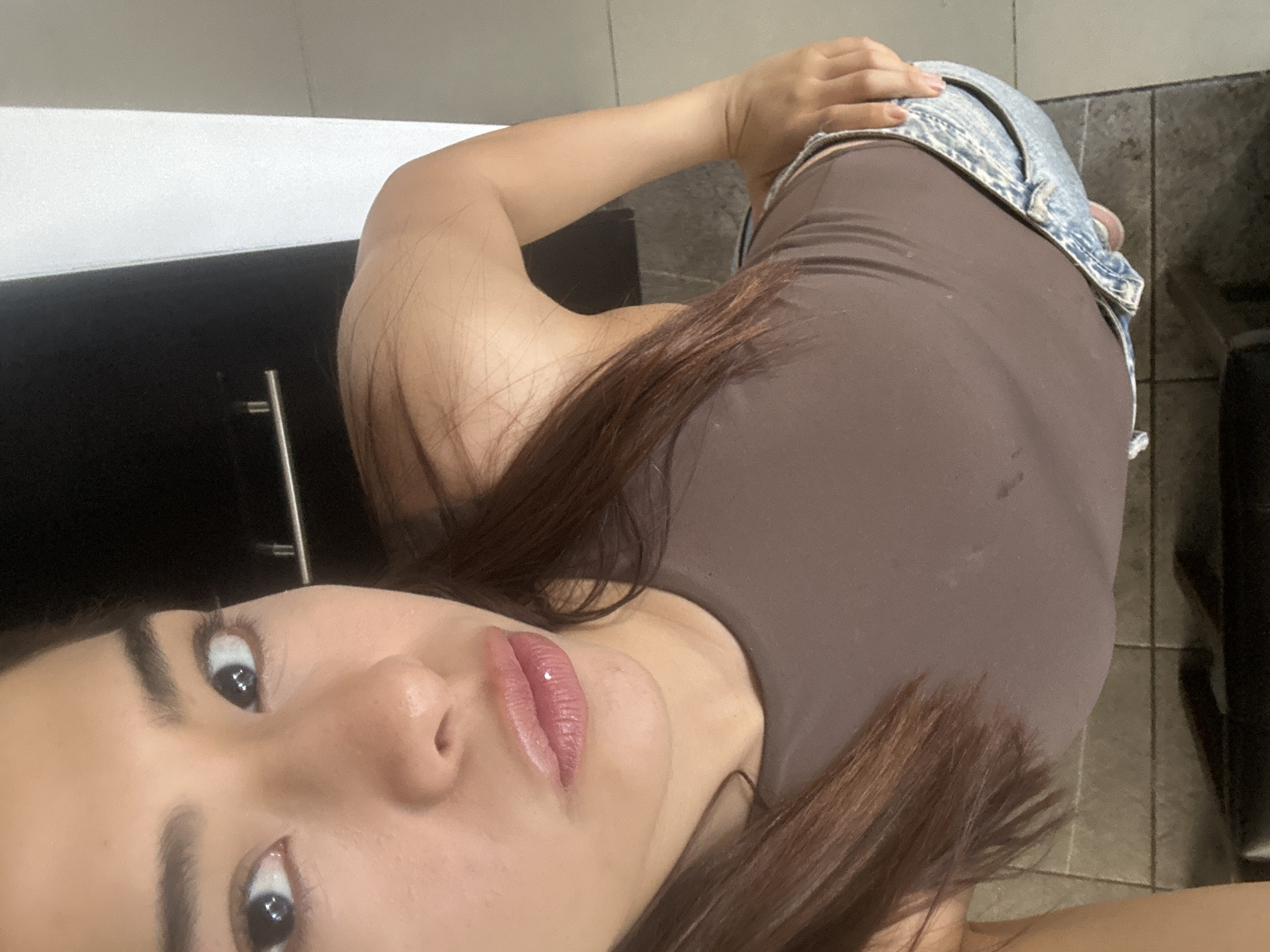An indoor, eye-level shot captures a woman lying down, looking directly at the camera with her head in the bottom left of the frame. The image is rotated 90 degrees clockwise. She has long, dark brown hair that cascades over her shoulder and chest. Her eyes are open, with visible eyeliner and mascara. Her lips are slightly parted, showing a glossy, pink-toned lipstick.
She is wearing a form-fitting, light brown or taupe sleeveless top or dress, paired with light-wash denim shorts or a skirt that is partly visible on her lower back. One of her hands rests on the denim garment.
The background consists of a dark, possibly wooden or laminate surface on the left, next to a white surface that could be a countertop or wall. A metal handle or bar is partially visible on the dark surface. The floor beneath her appears to be tiled in a dark, possibly grey or brown pattern. The lighting suggests an indoor setting, and no specific time of day is discernible. The overall atmosphere is casual and relaxed.
This scene is set in San Bernardino Tlaxcalancingo, Mexico. There is no visible text in the image.