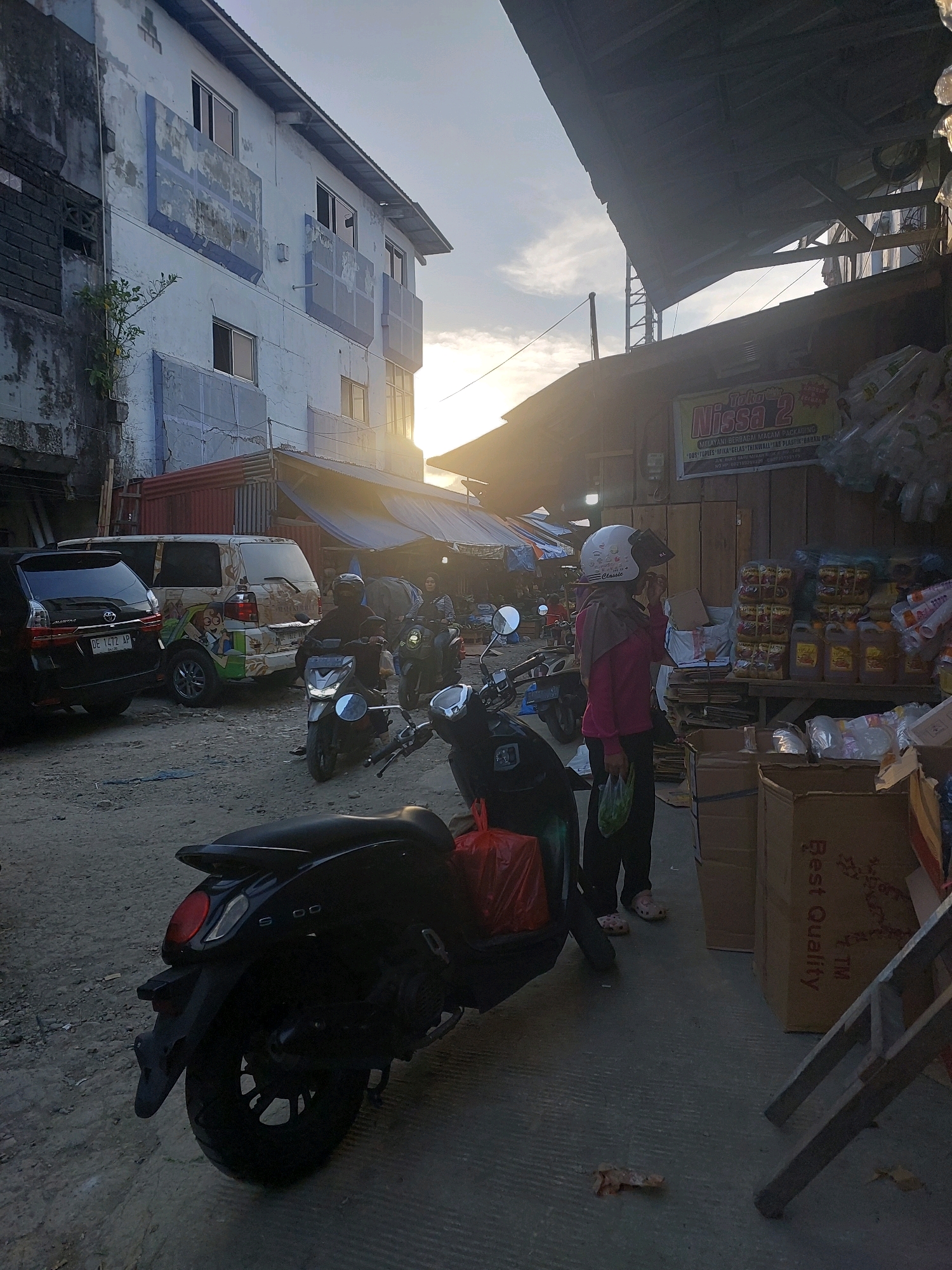 The image depicts an outdoor marketplace scene in Ambon, Indonesia, likely during the late afternoon or early evening due to the low sun angle and warm lighting. In the foreground, a black scooter is parked on a dirt and concrete surface, with a red bag on its seat. To the right, a woman wearing a pink shirt, dark pants, a grey hijab, and a white helmet is standing and holding a plastic bag. Behind her, a stall displays stacks of goods, including bottles of cooking oil and plastic packaging. A cardboard box with "Best Quality" printed on it is also visible.
In the mid-ground, a silver scooter is being ridden by a person wearing a dark helmet and jacket. To the left of this scooter, a white van with colorful cartoon graphics is parked, and further left, a black SUV is also parked. In the background, a multi-story building with a light blue facade and peeling paint stands, along with corrugated metal structures and awnings characteristic of a market environment. The sky is partly cloudy with the sun setting or low on the horizon, casting long shadows and creating a warm glow.