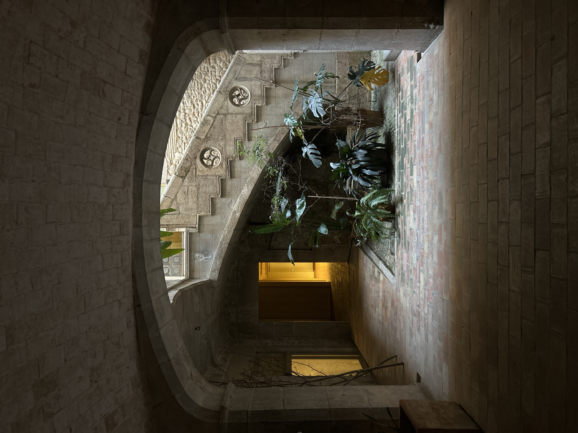 The image depicts an interior courtyard with a grand stone staircase, likely located in the historic old town of Girona, Spain. The courtyard is covered by a stone archway, with brick paving on the floor. Lush green plants are scattered throughout the space, with some placed in pots and others growing in a lower-level planter area. The staircase itself is made of stone, with decorative circular window-like openings along its side. The walls are constructed of rough-hewn stone blocks, giving the space an ancient and rustic feel. Natural light streams in from openings at the top of the courtyard and from a brightly lit doorway visible in the background. The overall impression is one of a well-preserved historical architectural space, possibly a private residence or a museum. There are no people visible in the image.