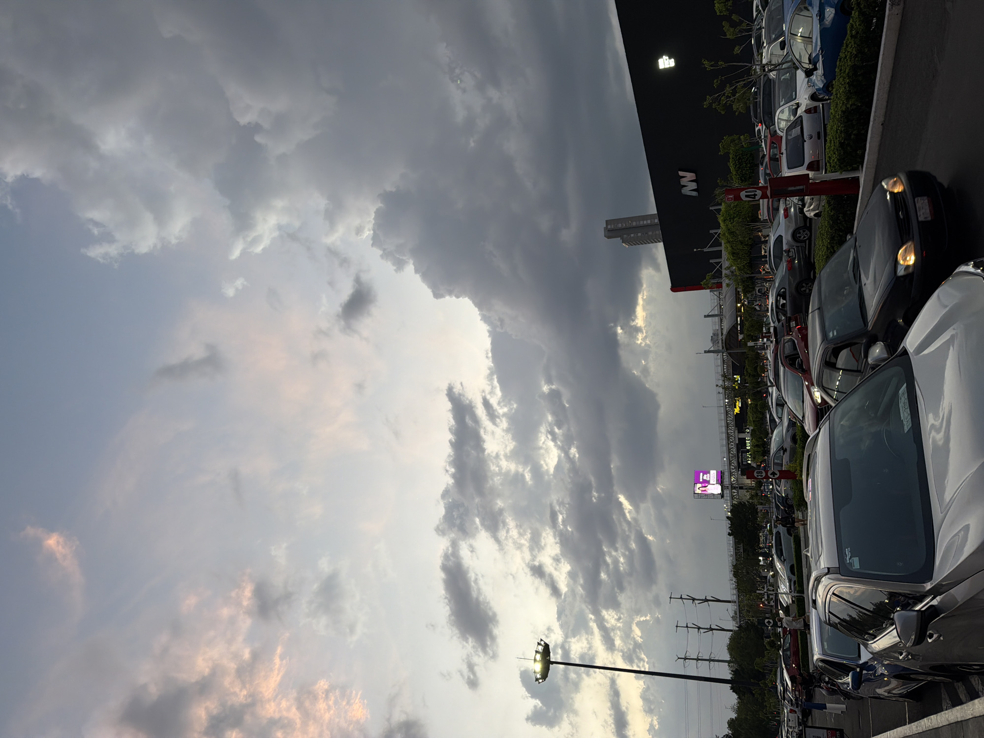 The image shows a parking lot in Puebla, Mexico under a cloudy sky. The parking lot is full of cars of various makes and models, densely parked in rows. Adjacent to the parking lot is a large retail building with a dark facade featuring a white "NW" logo. In the background, there are utility poles, trees, and a digital billboard displaying a person in pink. The sky is dominated by grey clouds, with some patches of lighter sky and a hint of pink near the horizon.