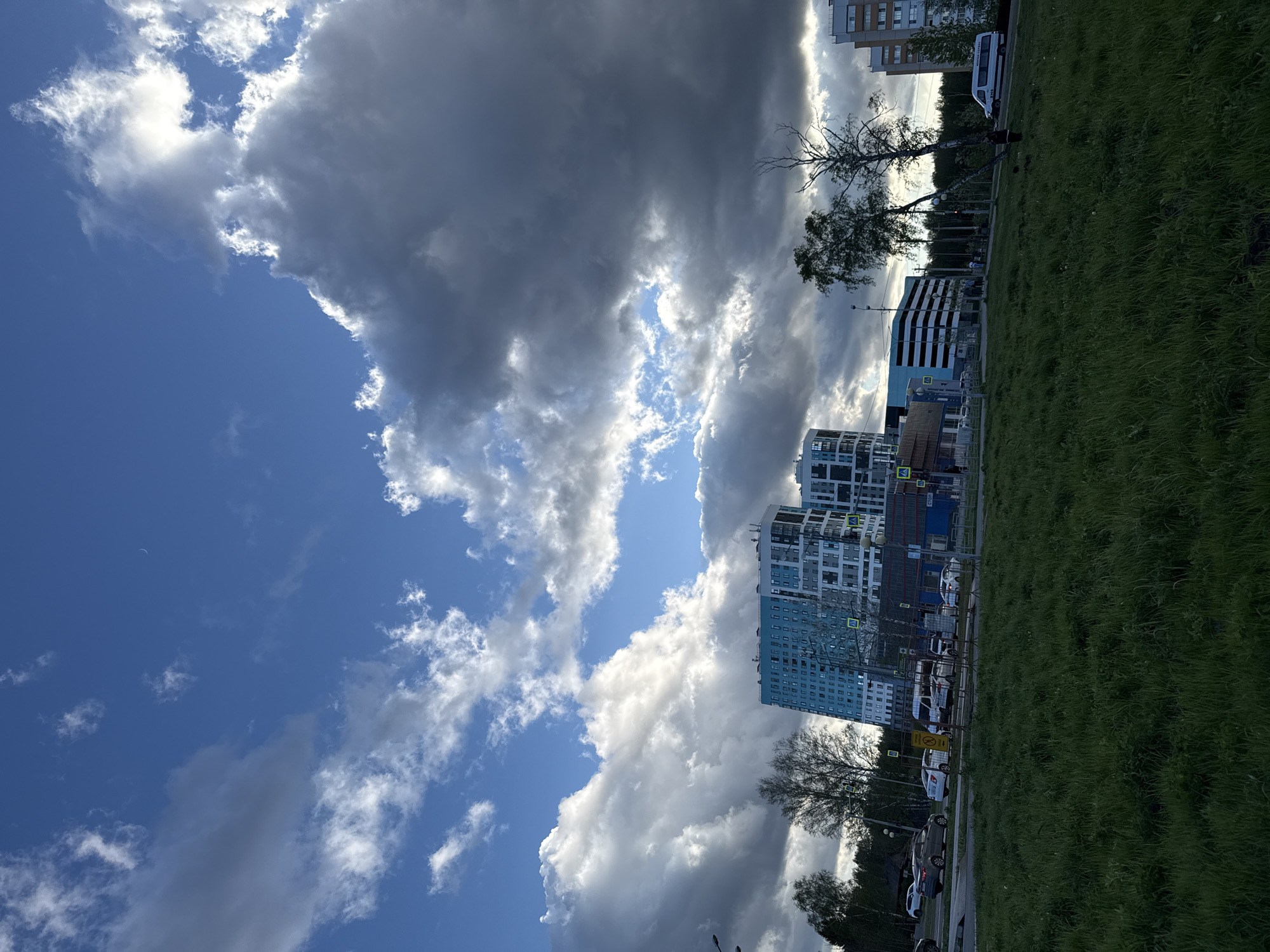 The image captures a vertical view of a landscape in Tobolsk, Russia. The sky dominates the upper half of the frame, showcasing a dynamic cloudscape with fluffy white and dark grey clouds against a backdrop of clear blue sky. A sliver of a crescent moon is barely visible amidst the blue sky. Below, urban elements are visible, including a building with a blue facade, characterized by numerous windows, along with other multi-story residential buildings. A strip of green grass fills the lower portion of the picture, bordering a street with parked cars and traffic signs. Scattered trees add vertical accents.
