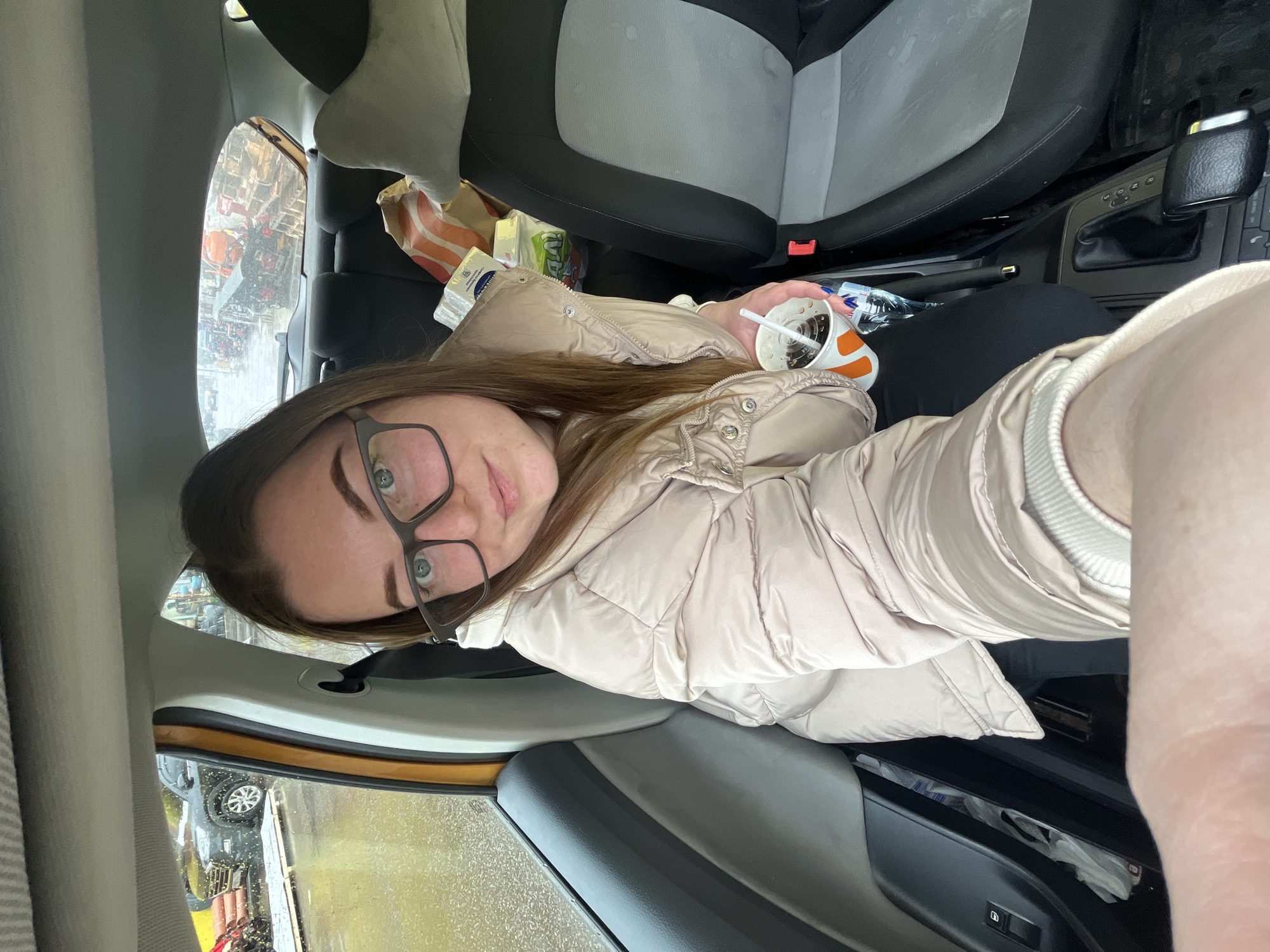 A woman is taking a selfie inside a car. She is wearing glasses and a light-colored puffer jacket, and she is holding a partially empty drink in her right hand. The interior of the car appears cluttered, with food wrappers and other items visible. Outside the car window, reflections suggest a street scene in Shevlyakovo, Russia.