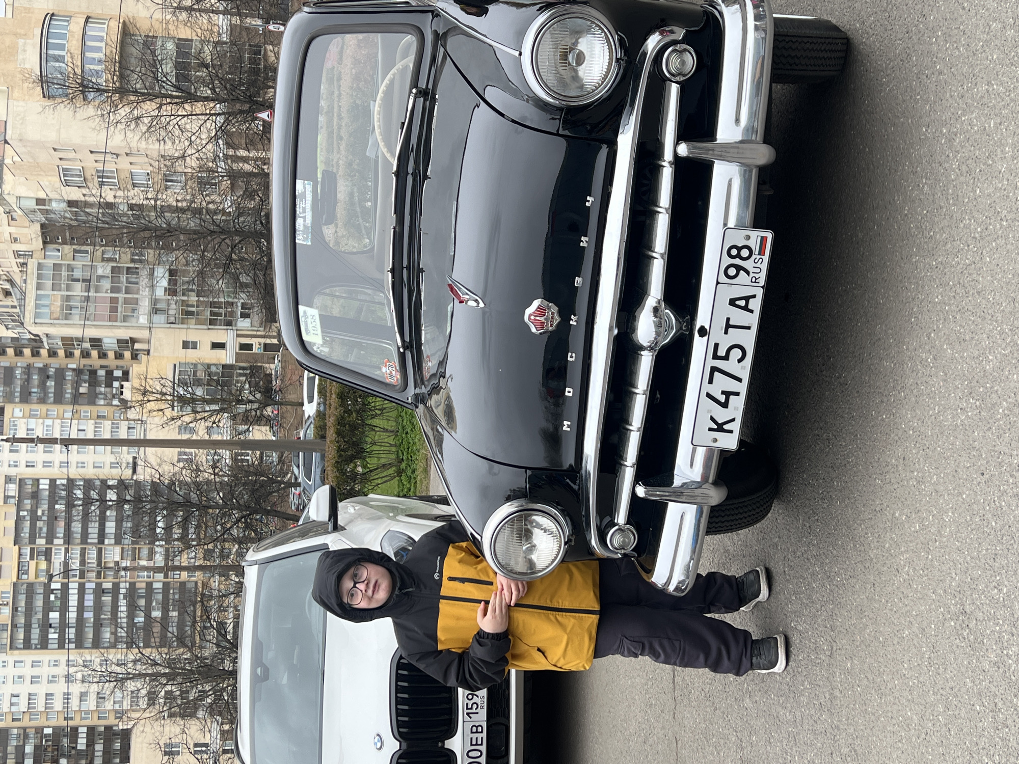 A black vintage car, possibly a Moskvich, is parked next to a white modern SUV in an urban setting, likely Saint Petersburg, Russia. A young person in a yellow and black jacket and glasses stands partially obscured by the black car. The car has a Russian license plate reading "K475TA98 RUS." A modern multi-story building is visible in the background, suggesting a residential or commercial area. The ground appears to be asphalt. The weather seems overcast. A Russian license plate on the white SUV is partially visible, with the digits "00EB159."