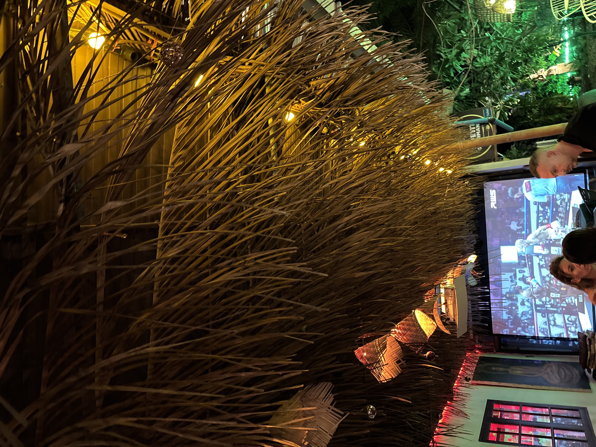 This image captures a vibrant evening scene in what appears to be an outdoor or semi-outdoor establishment, likely a bar or restaurant, in Baan Lamai, Thailand. The foreground is dominated by a decorative wall or roof covering made of densely arranged dried, straw-like natural materials, possibly palm fronds or bamboo strips. Numerous warm yellow string lights are intertwined within this material, casting a soft, inviting glow across the scene.
In the midground, several people are visible. A man with short hair is seen from the side, looking towards a large television screen. A woman's head is partially visible next to him, also observing the screen. The screen prominently displays a combat sport, appearing to be boxing or mixed martial arts, with two fighters in a ring and an audience in the background. The acronym "RWS" is visible in the lower part of the screen.
The setting incorporates various decorative elements, including elaborate woven lanterns or light fixtures hanging from the natural material, emitting an orange light. Small, reflective spherical ornaments are also present. In the upper right background, lush green foliage and trees are illuminated with subtle green lighting, contributing to a tropical ambiance. Below the television, a framed painting hangs on a wall, and a window with distinctive red-lit panes adds a striking color contrast. A sign behind the man has partially visible text, possibly "ETIT RMAN." The overall lighting and decor suggest a relaxed, engaging atmosphere at night.