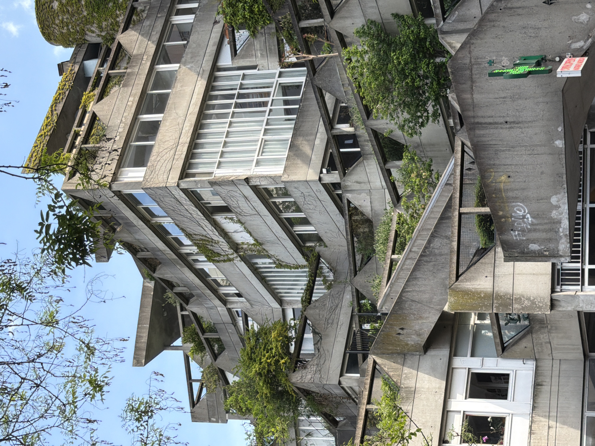 A photograph, rotated approximately 90 degrees clockwise, captures a striking brutalist-style concrete building in Ivry-sur-Seine, France, against a clear blue sky. The multi-story structure is defined by its complex, angular geometry, featuring numerous interconnected concrete beams, triangular sections, and recessed balconies. Lush green vegetation, including ivy and various bushes and small trees, extensively covers parts of the concrete facade and grows from planters within the building's design, creating a vibrant contrast with the raw grey material.
On the far left, a cylindrical, ivy-covered tower-like structure rises. The building is interspersed with numerous windows of varying sizes, predominantly featuring white frames, some of which are large and multi-paned. Tree branches with green foliage partially frame the scene from the lower left and upper right foreground.
Notable details include a green illuminated sign, possibly an arrow or directional symbol, attached to a concrete beam, alongside a red sign displaying "24H" vertically in white text. Faint graffiti markings are visible on the concrete in several areas. The bright daylight and clear sky suggest the photograph was taken during the day under fair weather conditions. The scene is static, with no people or discernible activity present.